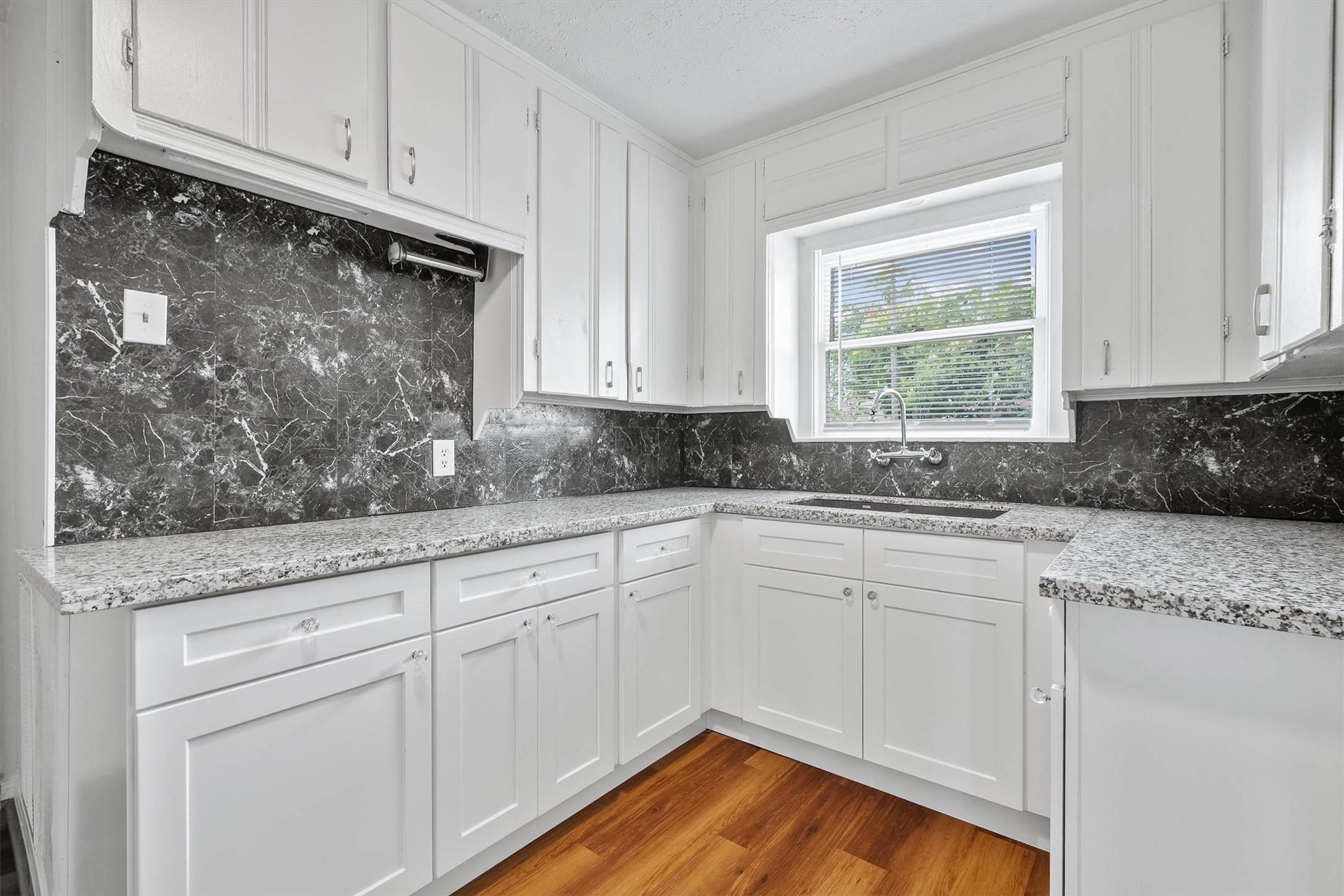 Bright kitchen featuring sleek white cabinetry, elegant granite countertops, and a striking marble backsplash, complemented by warm hardwood floors and ample natural light from the large window.
