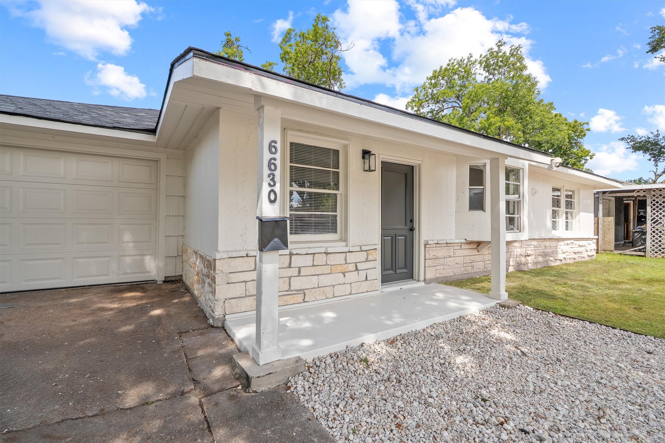Charming single-story home with a welcoming front porch, stone accents, and a spacious garage, set against a backdrop of lush greenery.