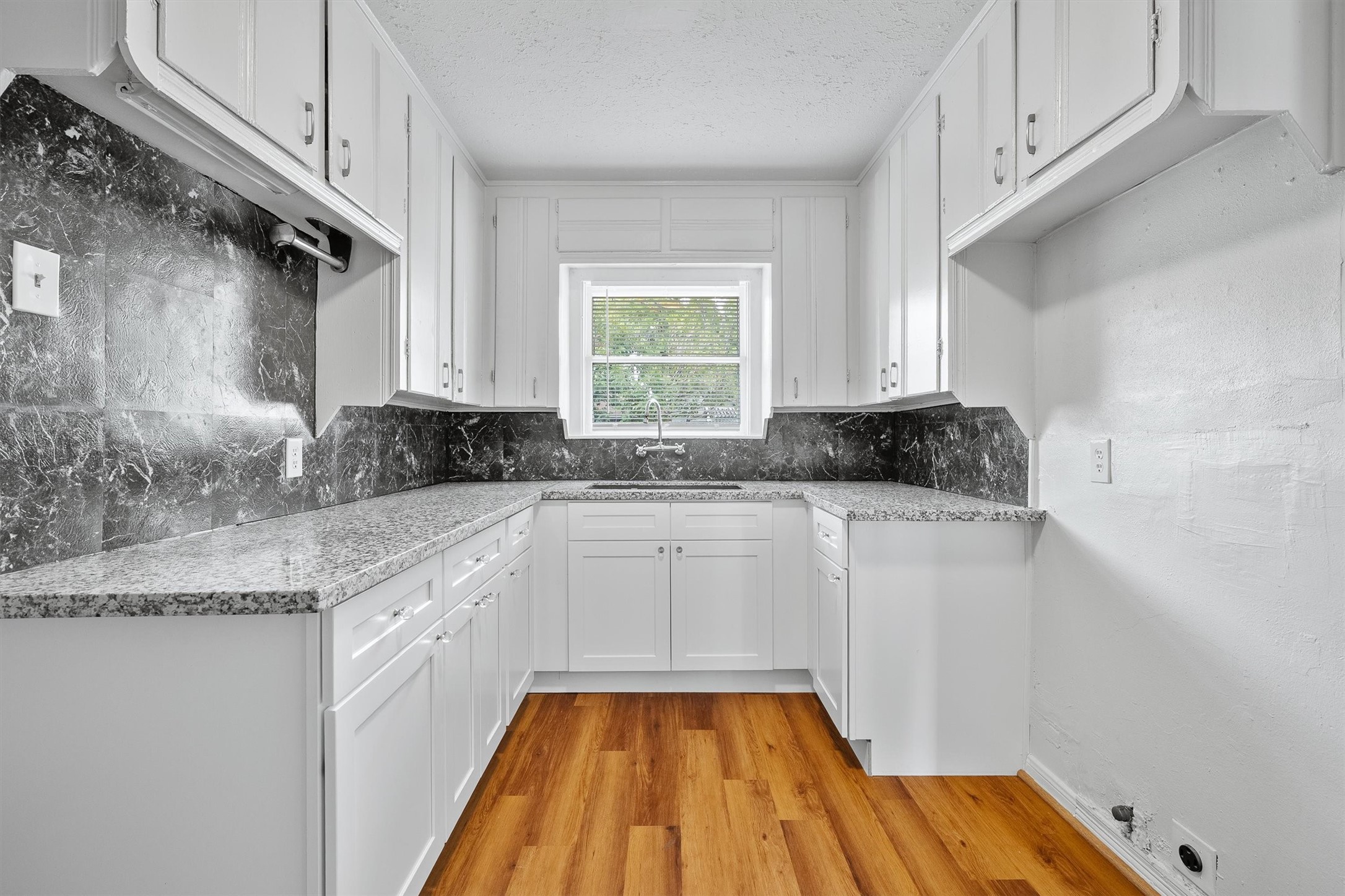 Bright kitchen with sleek granite countertops, classic white cabinetry, and a stylish black marble backsplash, complemented by warm wood flooring.