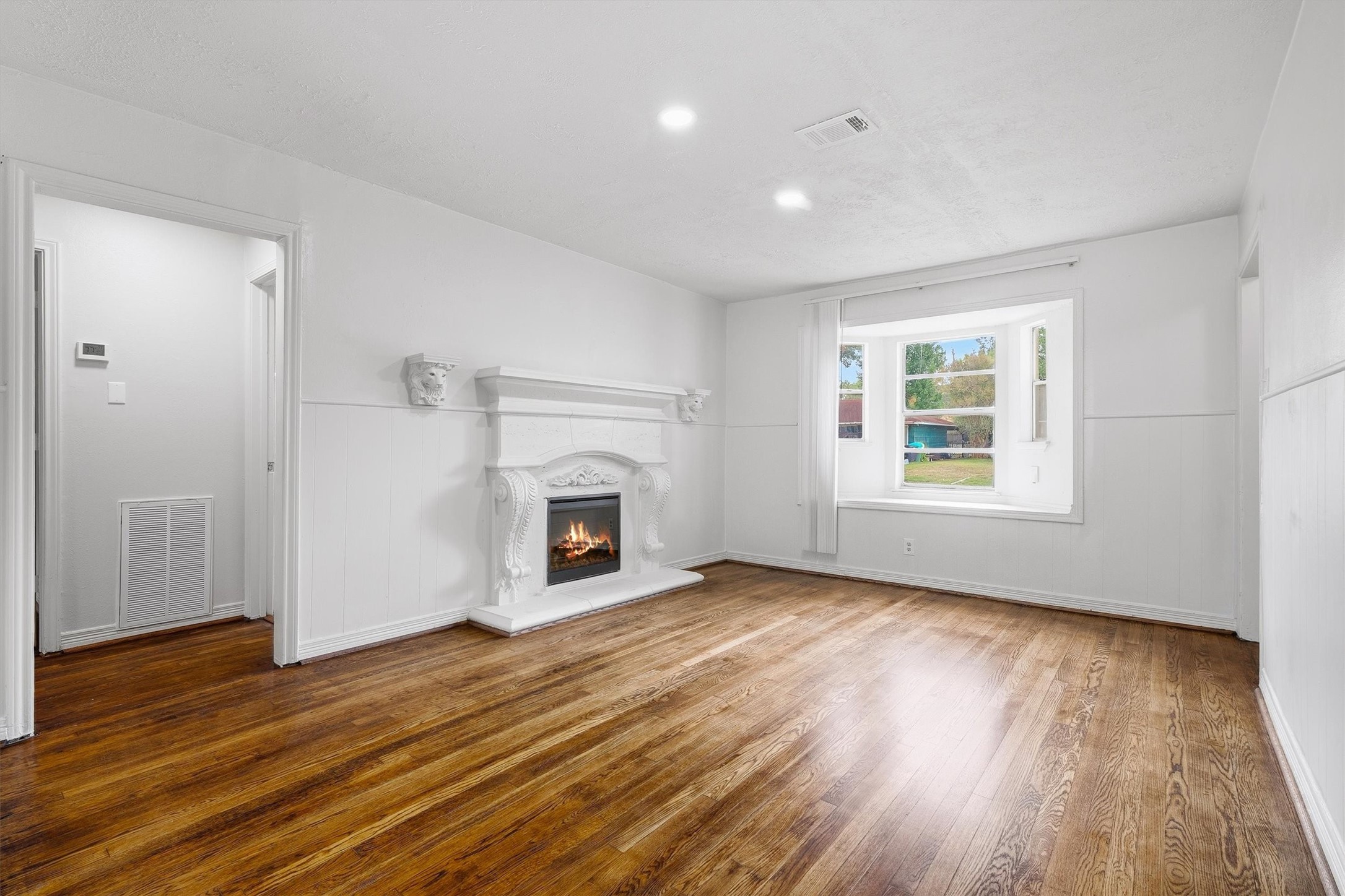 Charming living room with elegant white paneled walls, a decorative fireplace, and beautifully refinished hardwood floors, complemented by a large window that invites ample natural light.