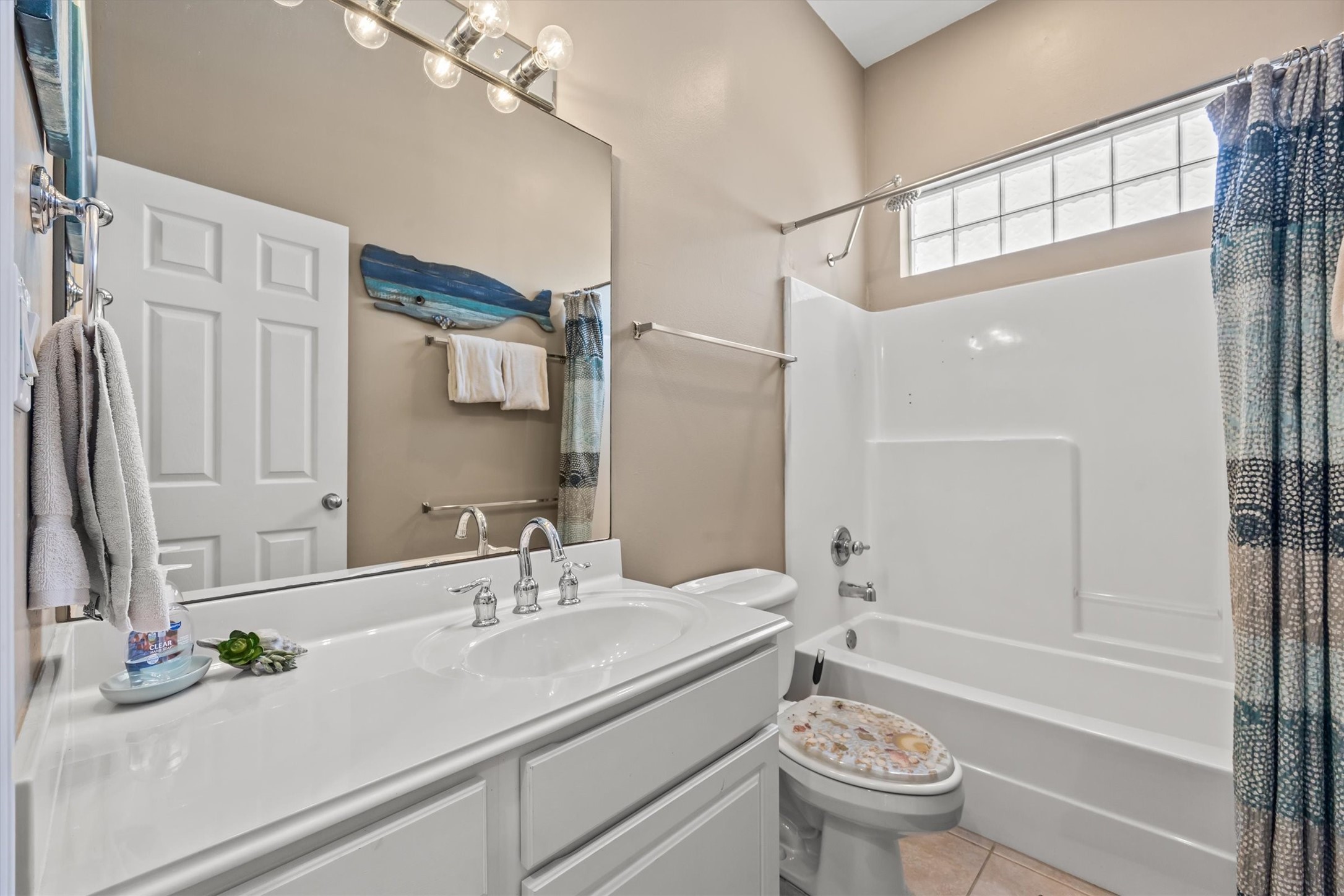 This bathroom features a clean, modern design with a large mirror, ample lighting, and a white vanity. The space includes a bathtub with a shower, accented by a colorful curtain. Natural light enters through a glass block window, enhancing the room's bright and airy feel.