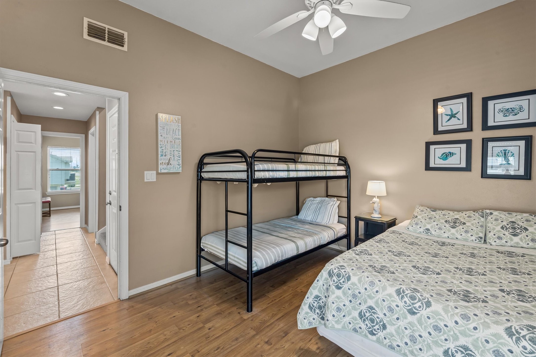 This cozy bedroom features a bunk bed and a queen-sized bed, with a ceiling fan for comfort. The space is warmly painted and decorated with coastal framed art. A hallway leads to an additional bedroom.
