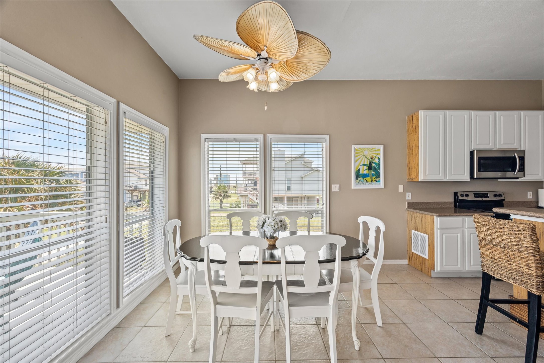 Bright dining area with large windows offering ample natural light. Features a large table with white chairs for family groups, a unique ceiling fan, and a view of a sunny day or a beautiful sunset. Conveniently connected to the modern kitchen with white cabinetry and stainless steel appliances.
