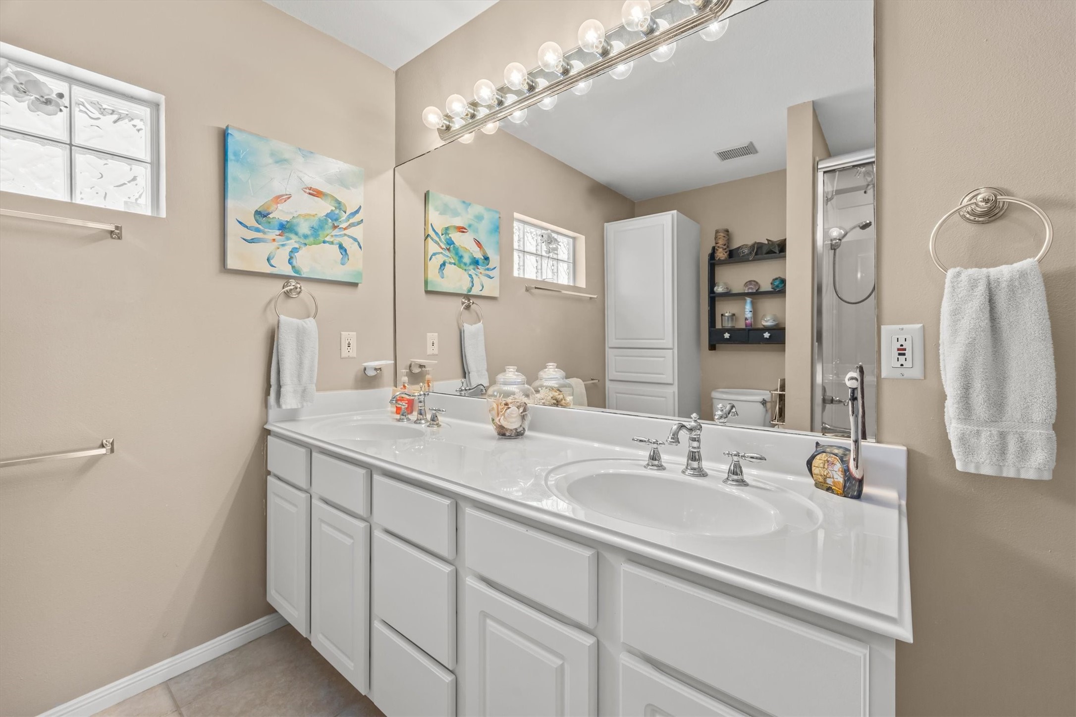 This bathroom features a double vanity with sleek white cabinetry, a large mirror with great lighting, and neutral-toned walls. It includes decorative artwork, a glass block window for natural light, and a shower area. The space is modern and inviting.