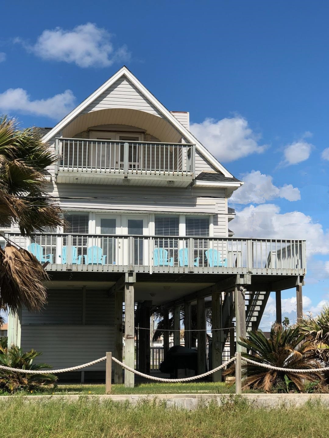 This is a charming elevated beach house with a spacious deck, perfect for enjoying ocean views. It features two levels of outdoor seating, palm trees, and a light, airy design. Ideal for coastal living.