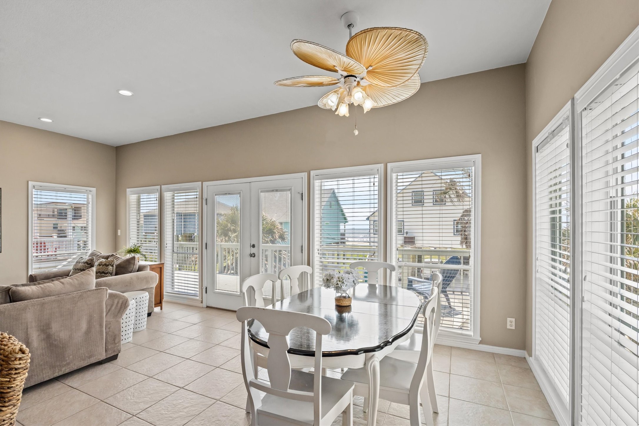 This bright and airy living space features large windows with white blinds, a stylish ceiling fan, and an open layout that includes a cozy sitting area and a dining table. The space opens to the deck with views of sunny blue sky and the beach offering a welcoming and comfortable atmosphere.