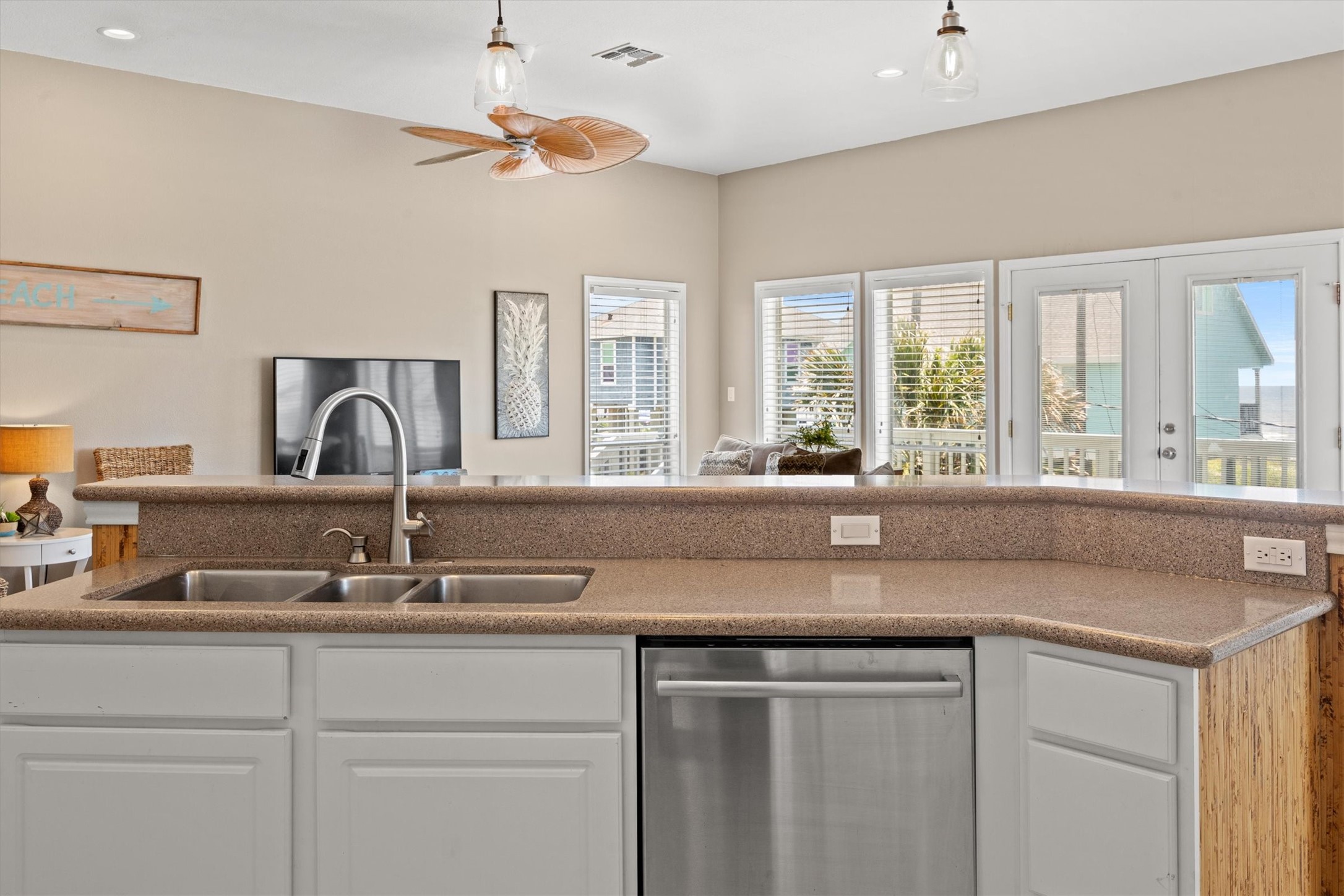 This photo shows bright, open kitchen overlooking the living area. There's a modern kitchen island with a double sink and stainless steel dishwasher backed by the roomy bar for casual eating. The space is well-lit with natural light from large windows and glass doors that lead to an outdoor area. The room features neutral tones and a ceiling fan, creating a welcoming atmosphere.