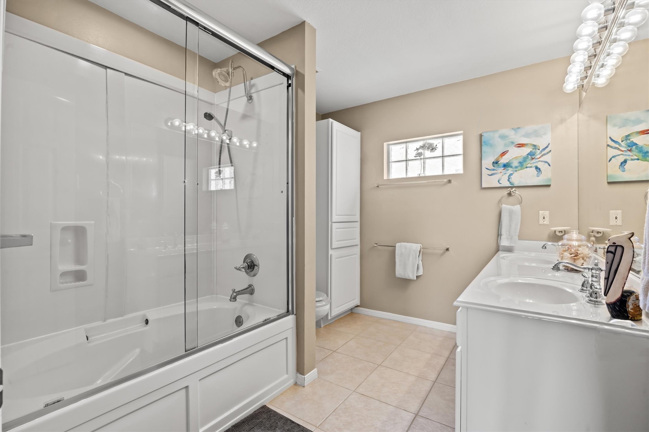 This clean, modern design includes a glass-enclosed shower-tub combo, a double-sink vanity with ample counter space, and neutral tile flooring. Bright lighting and a small window enhance the airy feel.