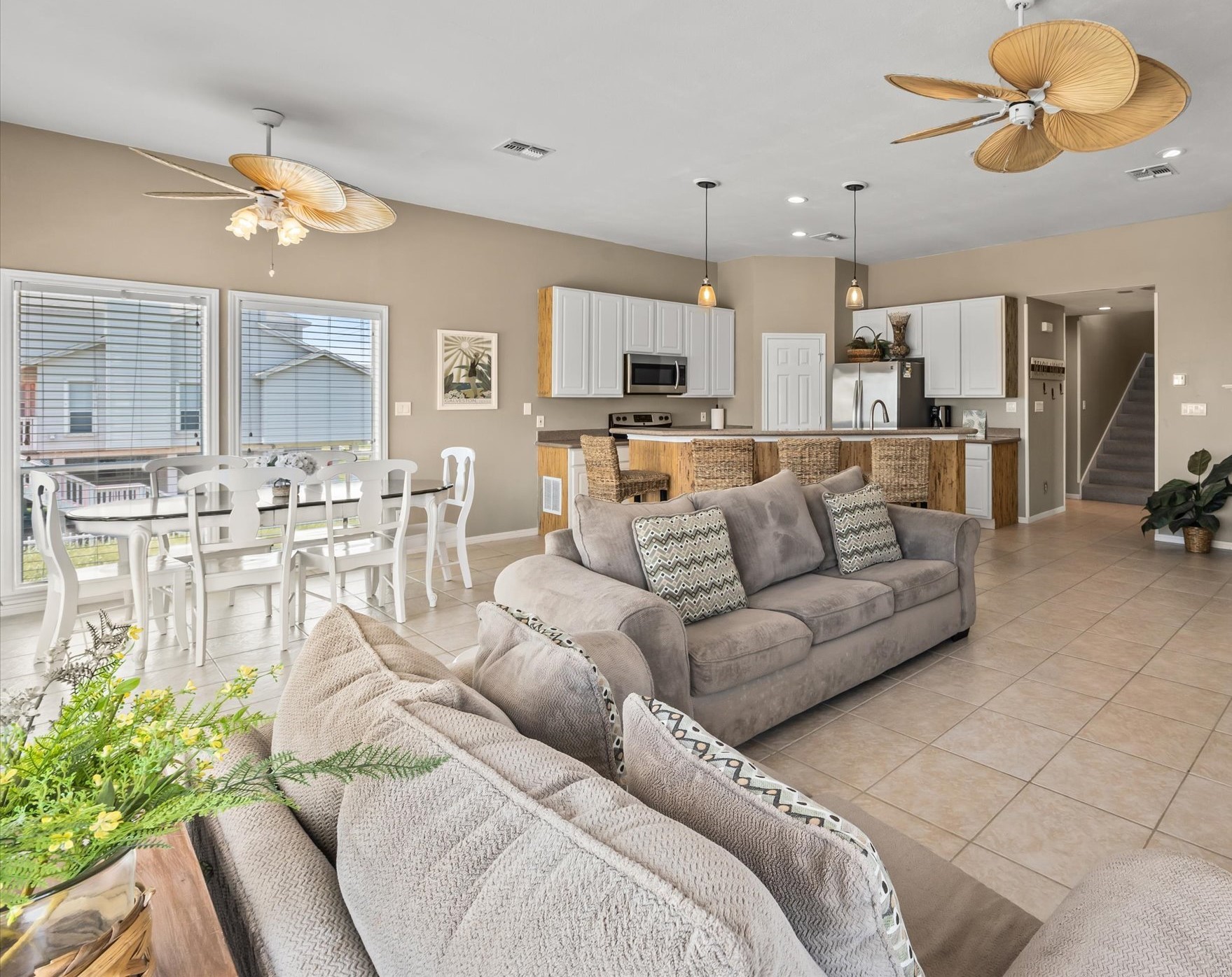 This open-concept living space features a modern kitchen with stainless steel appliances and a breakfast bar, a cozy dining area with natural light, and a comfortable lounge area. The room is adorned with stylish ceiling fans and has a neutral color palette, creating a warm and inviting atmosphere.