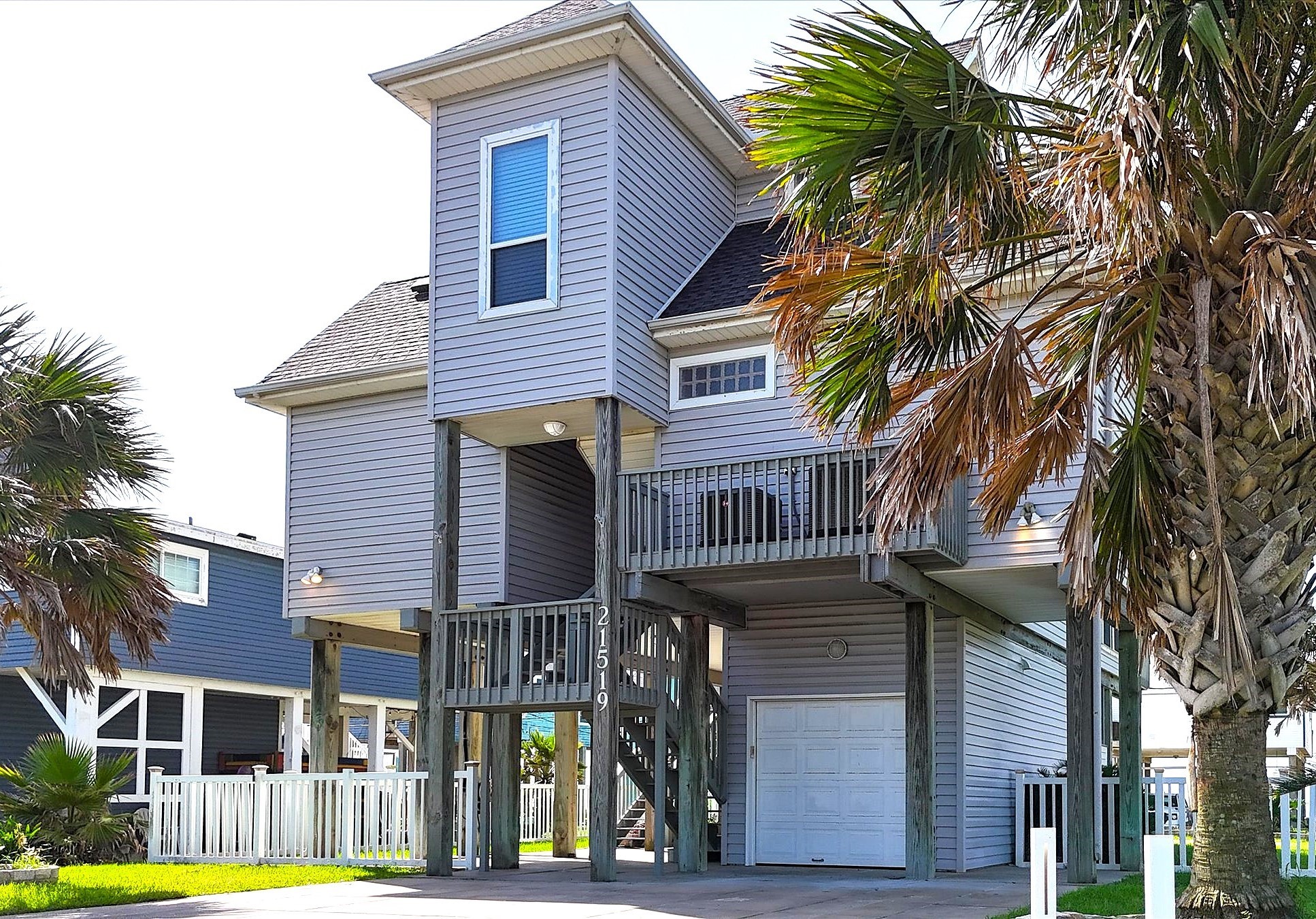 This photo shows the underside of the raised coastal home with a wooden staircase leading to the main floor, surrounded by a grassy yard and palm trees. The space beneath the house is open with concrete flooring, providing ample parking or storage.