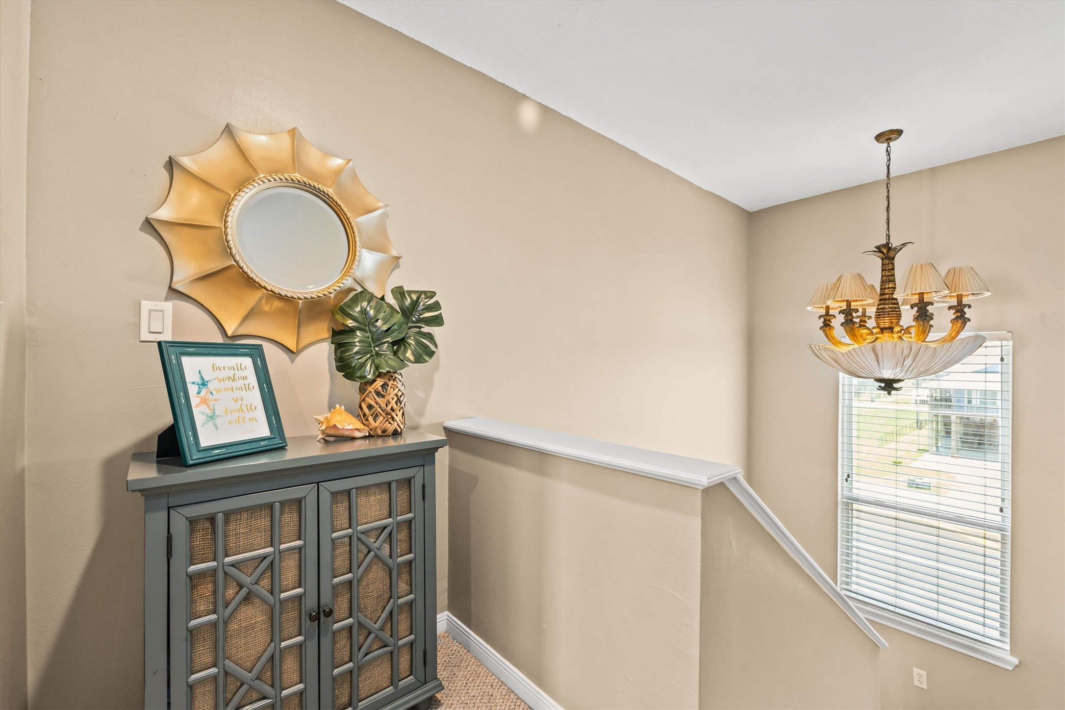 This photo shows an open hallway landing outside of the primary suite featuring a cabinet topped. A stylish sunburst mirror hangs above, and a classic chandelier illuminates the space. A window with blinds provides great natural light.