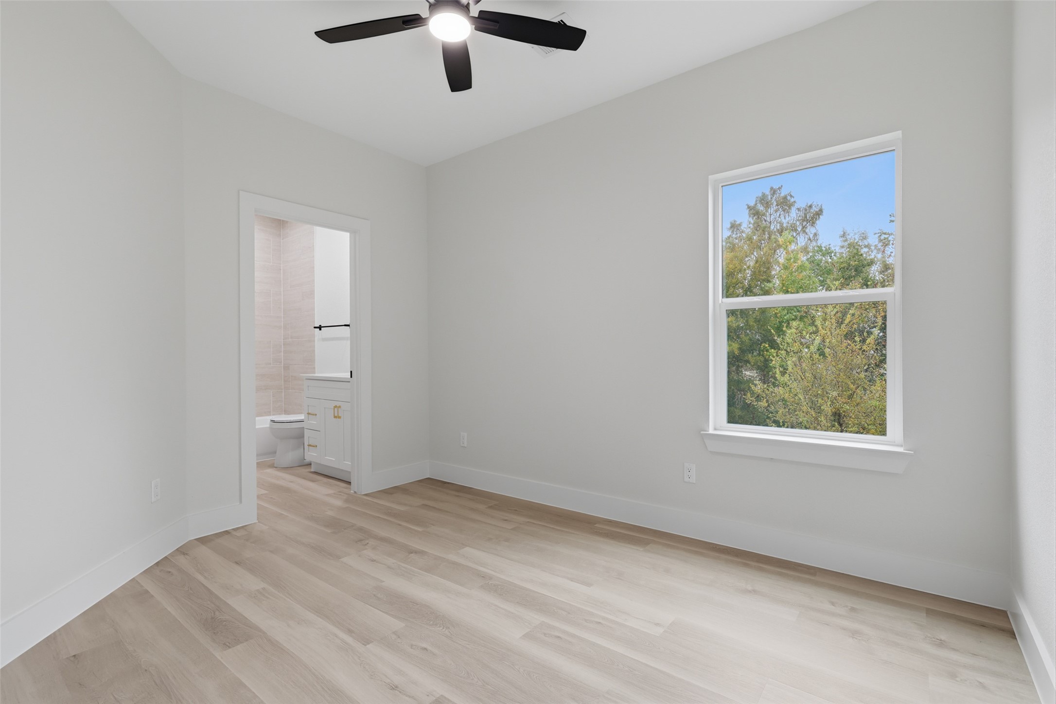 Bright and airy bedroom featuring sleek LVP floors, a ceiling fan, and a large window with a view of lush greenery, leading to a modern en-suite bathroom.