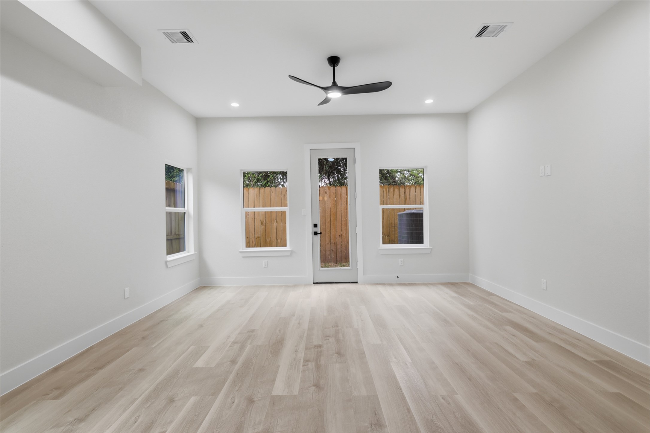 Bright and spacious living room featuring sleek wood flooring, recessed lighting, and a modern ceiling fan, with direct access to a fenced backyard through a glass door.