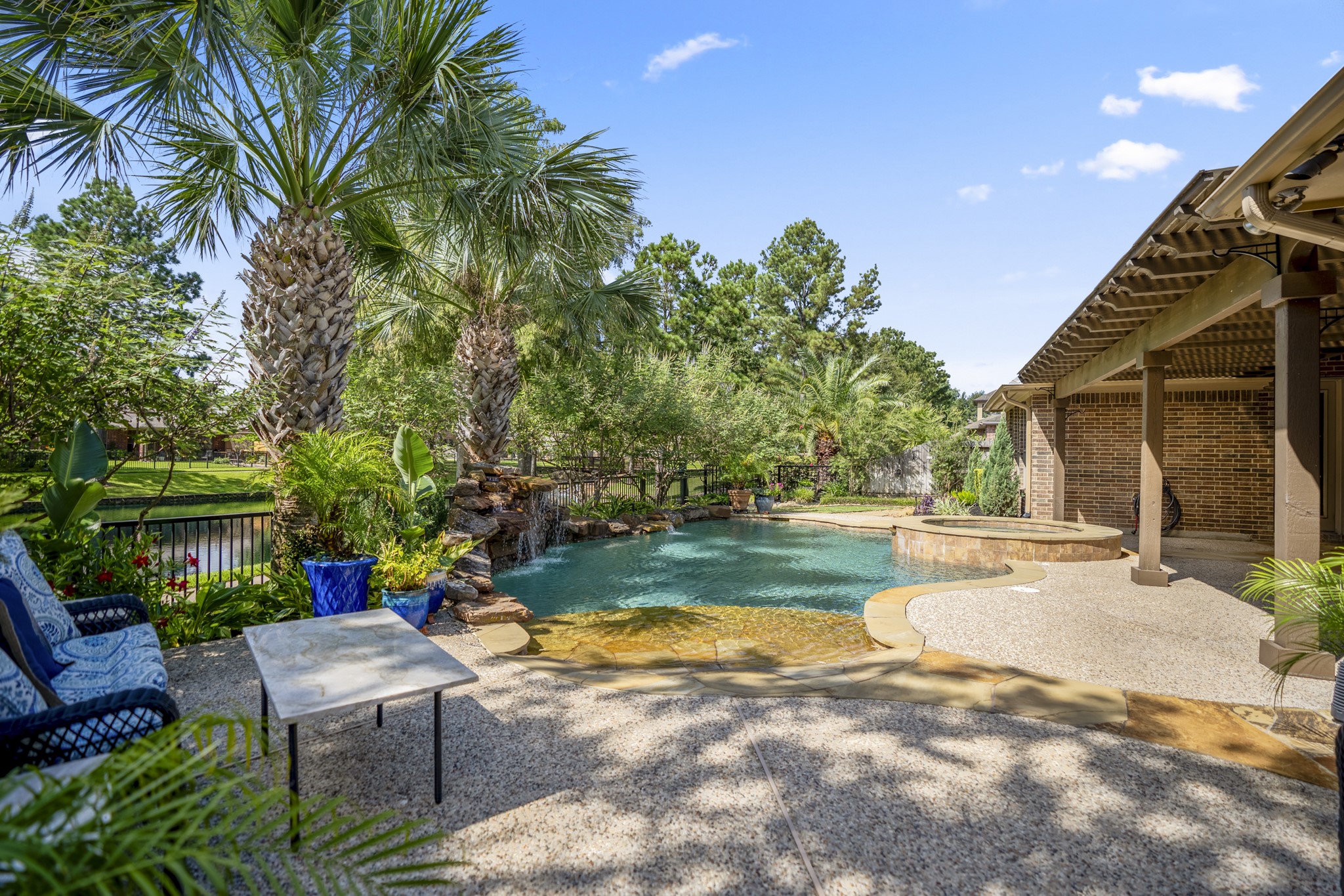 Poolside from the far end of the backyard, this view captures the beach-style pool entry, raised spa, and water beyond the yard for a fuller sense of the outdoor setting. The covered pergola stretches along the right side of the home, while palm trees and layered landscaping wrap the space with a relaxed, resort-style feel made for slow evenings, weekend swims, and time outside.
