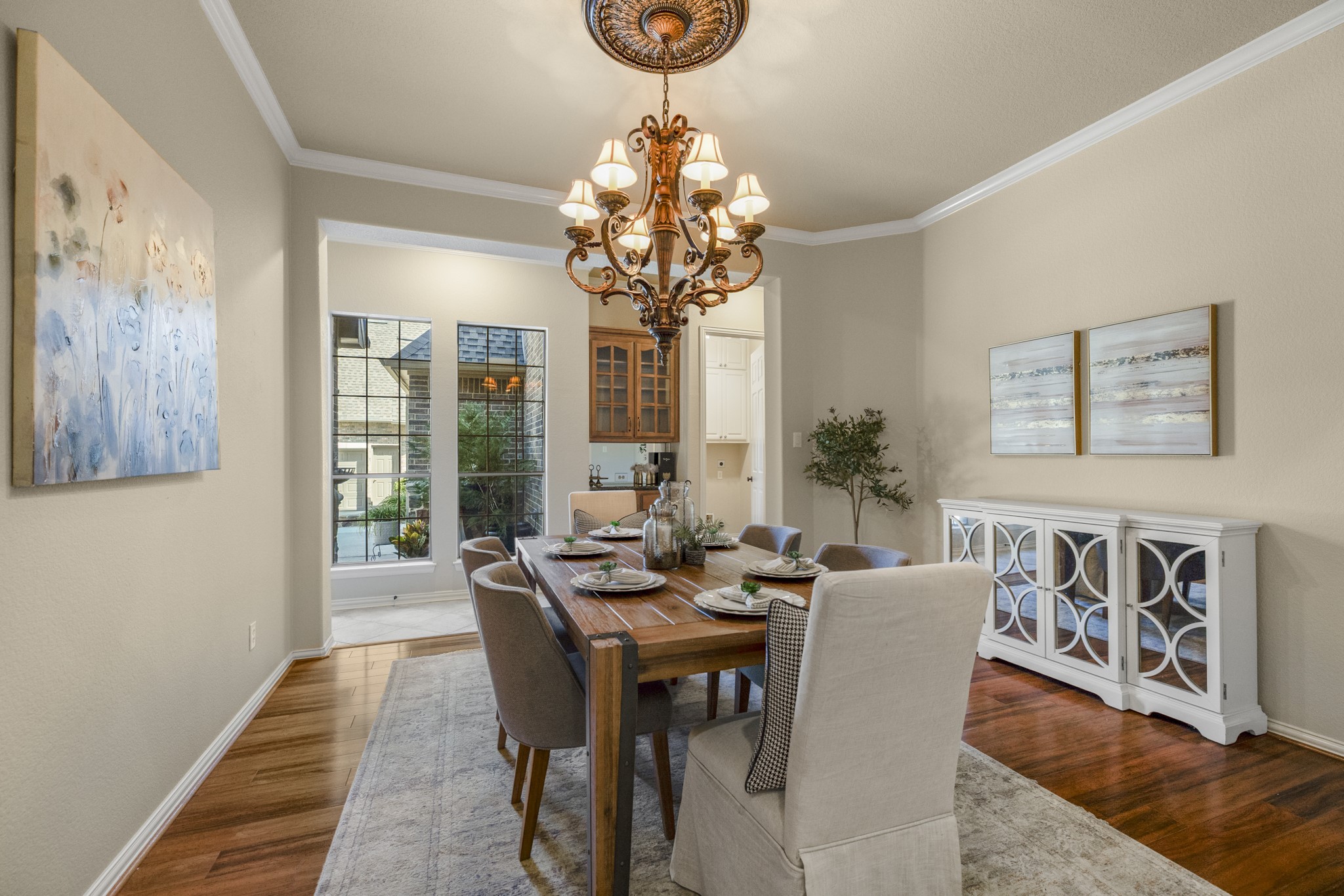 Standing at the foyer, the formal dining room feels elevated and intentional with mahogany wood floors, crown molding, and a detailed chandelier with a ceiling medallion overhead. The room offers space for a full table setting plus a buffet or display piece along the wall, while the wide opening ahead keeps it connected to the kitchen and breakfast area. Just beyond, the built-in dry bar/butlers pantry area adds a polished transition, with the utility room door tucked along that back wall.