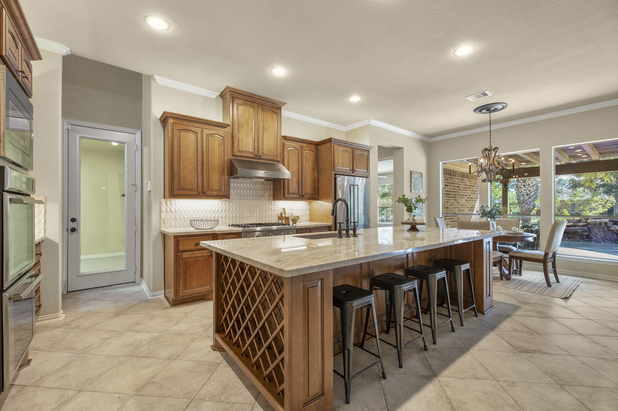 Inside the kitchen, the homes custom finish really comes together with Taj Mahal quartzite, Benedettini cabinetry, a Dal Tile backsplash, undercabinet lighting, and island seating that keeps the space warm and connected. Built-in wine storage adds a thoughtful detail at the end of the island, while the glass door on the left leads to the separate third garage bay and the breakfast nook ahead opens the view toward the covered patio, pool, and water-front backdrop beyond.