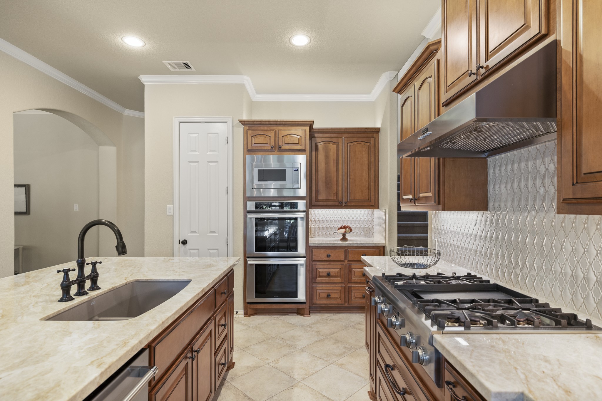 Along the working side of the kitchen, Taj Mahal quartzite, rich cabinetry, stainless appliances, and the upgraded tile backsplash keep the custom finish consistent from every angle. The refrigerator lines the right side, while the door on the left opens to the pantry, keeping storage close to the prep and cooking areas without interrupting the flow back toward the island.