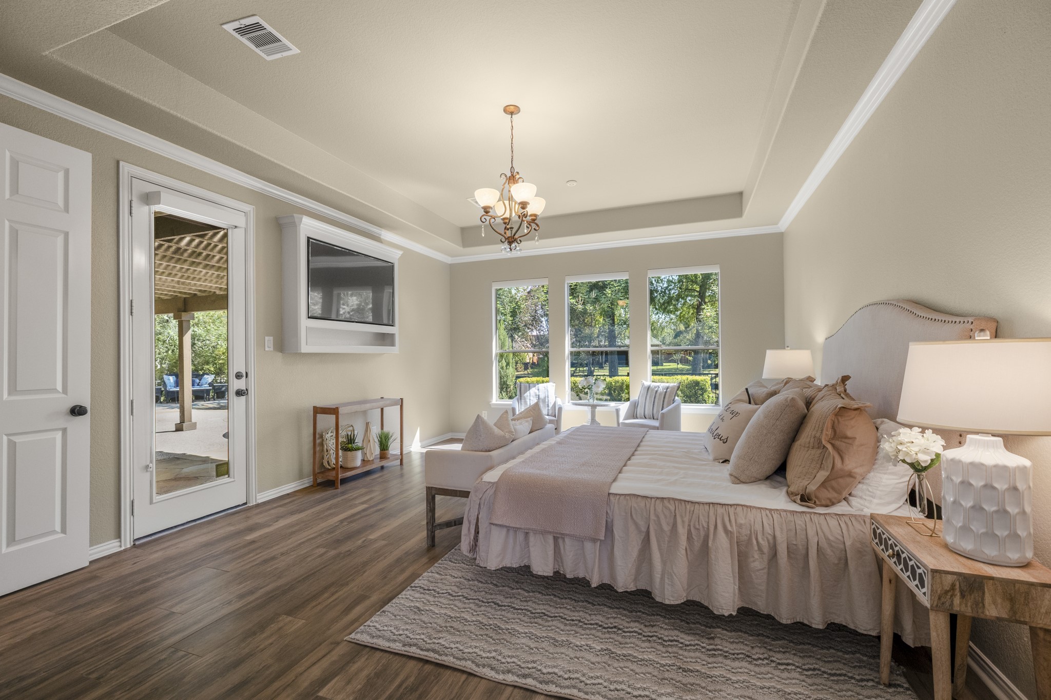From this angle, the primary suite feels closely connected to the outdoor living spaces, with a patio door on the left of the TV and rear windows bringing in views toward the pool, greenery, and water beyond. The TV built-in along the wall, tray ceiling, crown molding, and wide-plank wood-look tile add to the rooms custom finish, creating a relaxed retreat before the walkthrough continues into the remodeled primary bath.