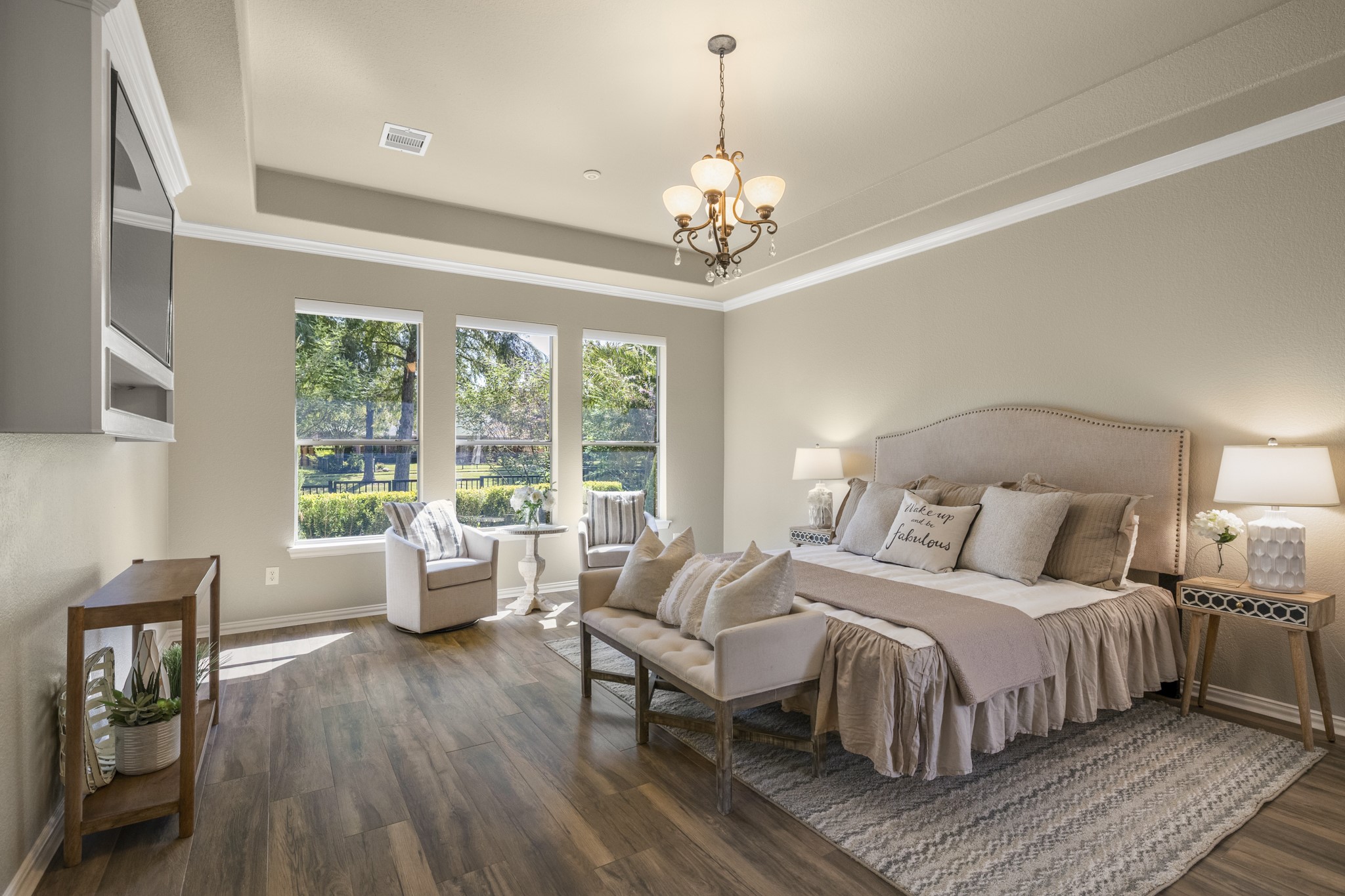 From the living room, the walkthrough moves into the primary suite, where rear windows bring in natural light and a glimpse toward the outdoor living area beyond. Crown molding, a tray ceiling, and wide-plank wood-look tile give the bedroom a refined, relaxed feel, while its location just off the living room keeps the suite connected to the homes water-view lifestyle before continuing into the remodeled primary bath.