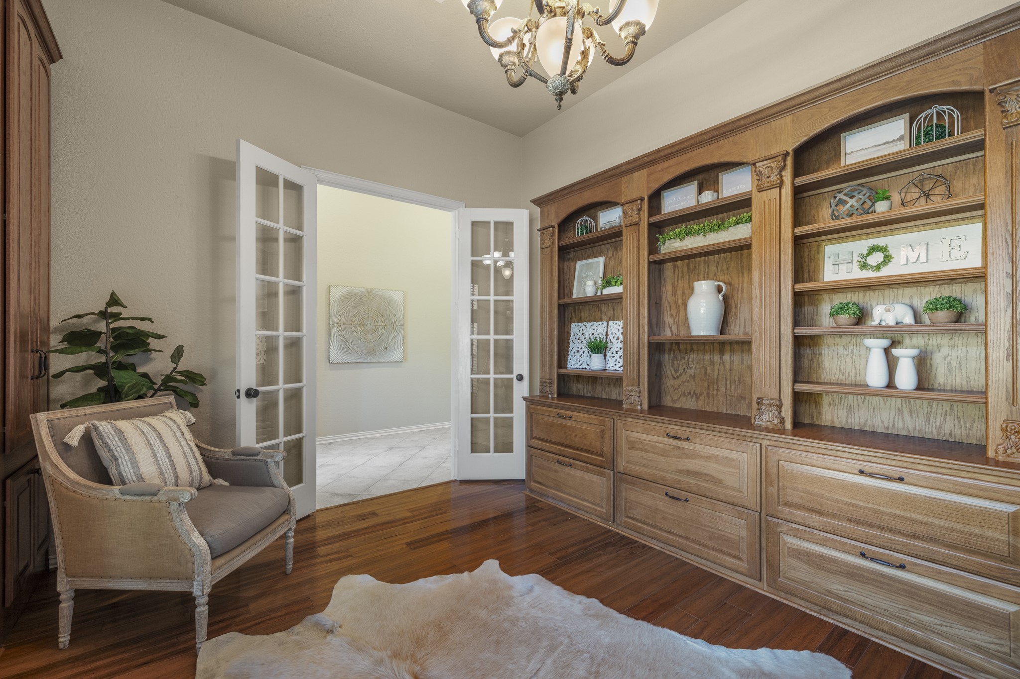 From this angle, the studys craftsmanship takes center stage, with richly detailed mahogany cabinetry, glass-front display cabinets, open shelving, and beautifully finished trim creating a sophisticated library atmosphere. Mahogany wood flooring and an elegant chandelier add warmth, while the French doors connect the space to the foyer, offering an inviting setting for working from home or settling in with a good book.