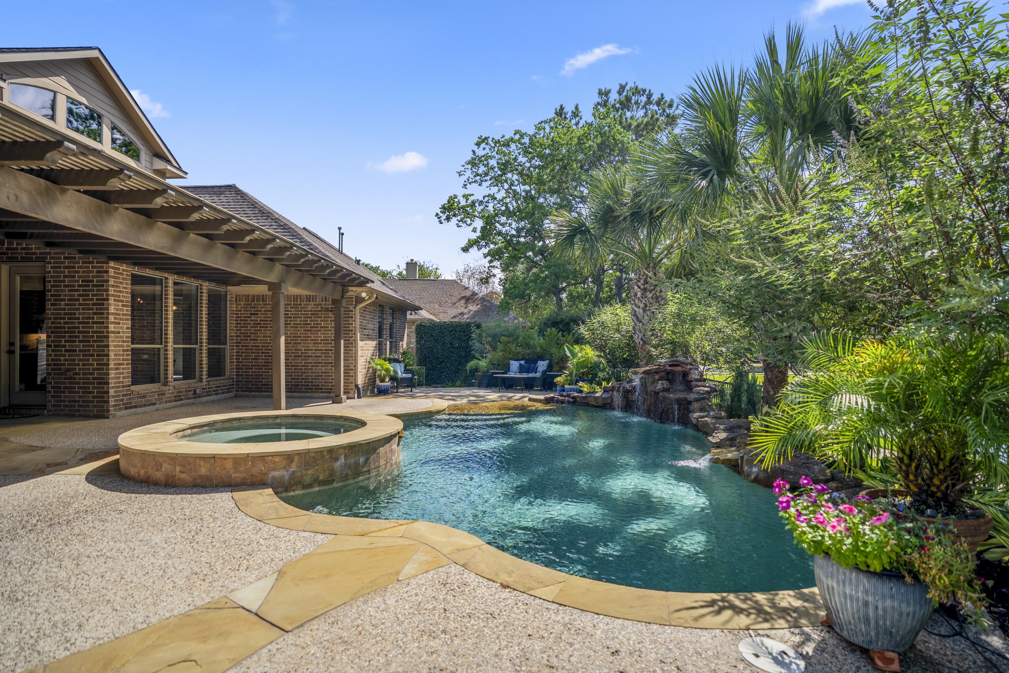 Poolside, the backyard feels wrapped in greenery, with the raised spa, curved pool, and surrounding landscaping creating a private-feeling place to relax outdoors. The covered pergola stretches along the back of the home on the left, while palm trees and layered shrubs soften the view and set up the kind of evening backdrop the sellers loved most.