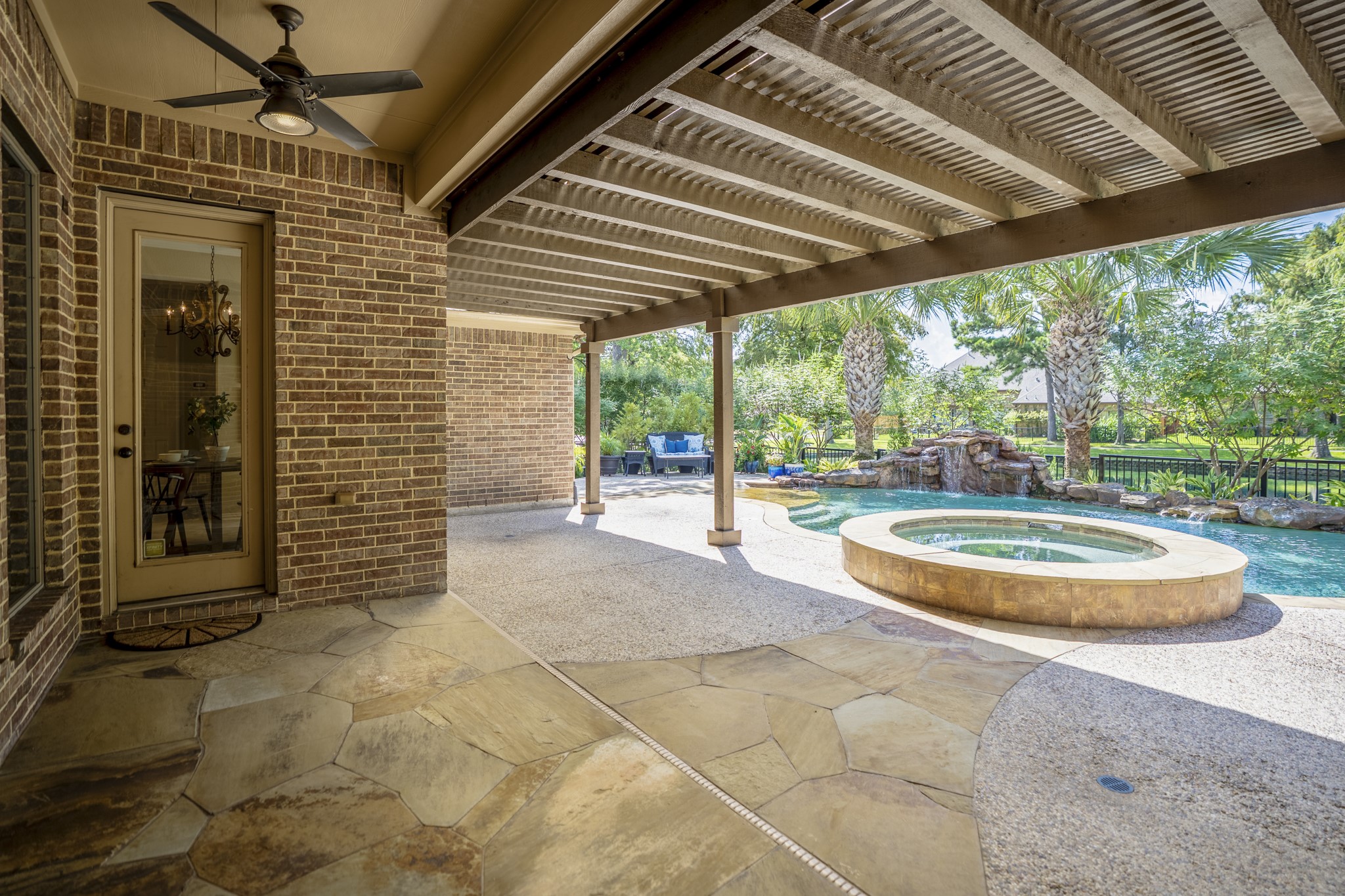 Beneath the 30 covered pergola, the patio becomes an outdoor room made for more than sunny afternoons, with stone underfoot, support columns, and shade stretching across the back of the home. The raised spa sits just beyond the covered area, keeping the pool and water-view setting close while giving this part of the backyard a relaxed, lounge-like feel.