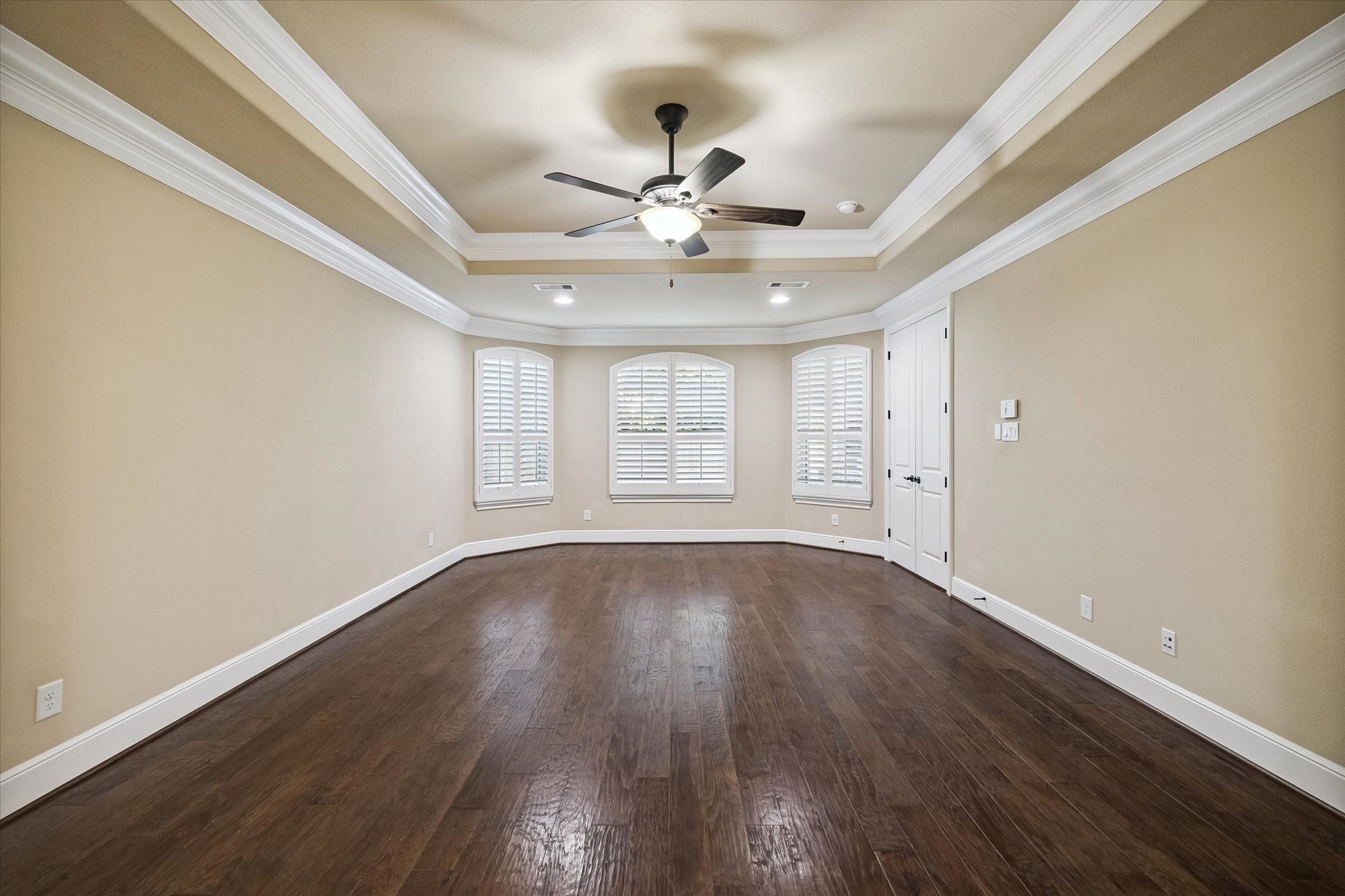 The generously sized primary suite offers a peaceful retreat with rich hardwood flooring, an elegant tray ceiling accented by crown molding, and a spacious sitting area surrounded by plantation-shuttered windows that fill the room with natural light.