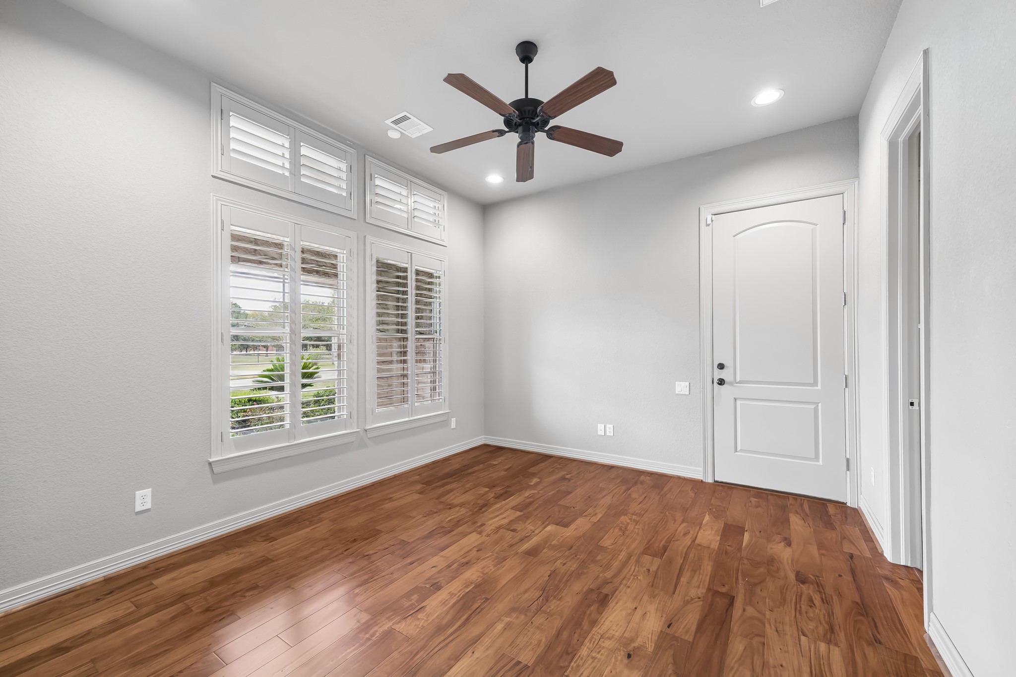 Bright and airy room featuring rich hardwood floors, a ceiling fan, and large windows with plantation shutters, perfect for natural light and ventilation.  Office or guest room with access to the garage with half bath.
