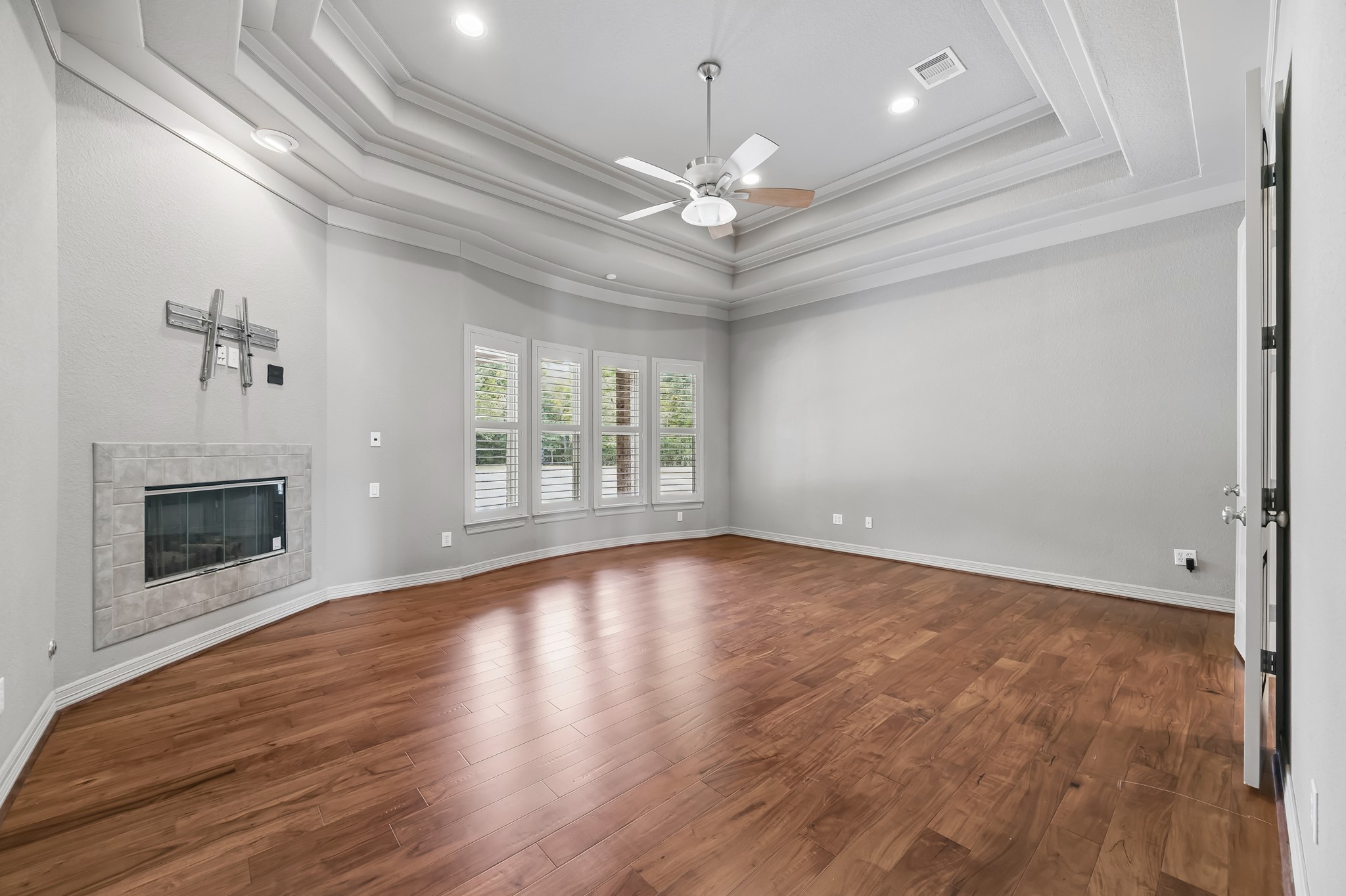 Spacious Primary Bedroom with elegant tray ceiling, rich hardwood floors, and a cozy modern fireplace, perfect for relaxation.