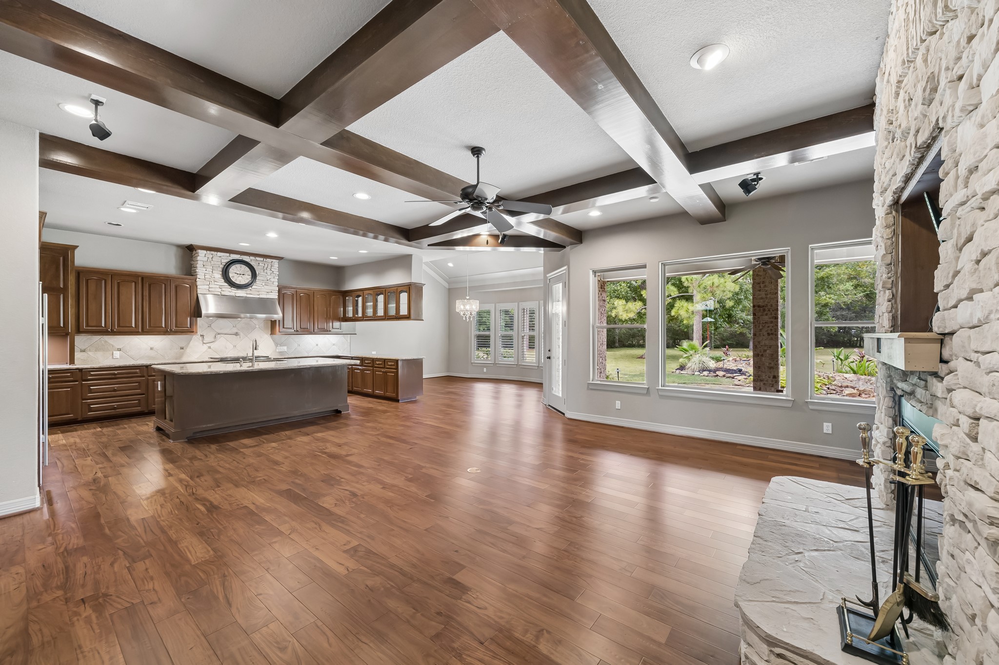 Spacious open-concept living area featuring elegant wood flooring, exposed ceiling beams, and a cozy stone fireplace, seamlessly flowing into the modern kitchen with rich wood cabinetry and a large island. Expansive windows offer stunning views of the lush backyard.