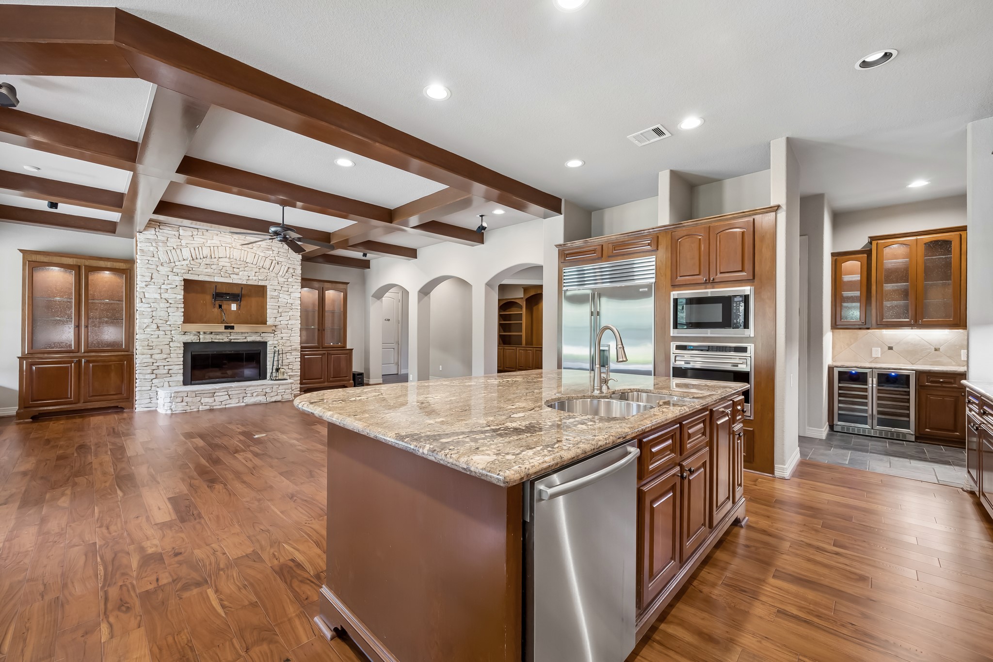 Spacious open-concept kitchen and living area featuring elegant wood-beamed ceilings, a large stone fireplace, and a central island with granite countertops, perfect for entertaining.