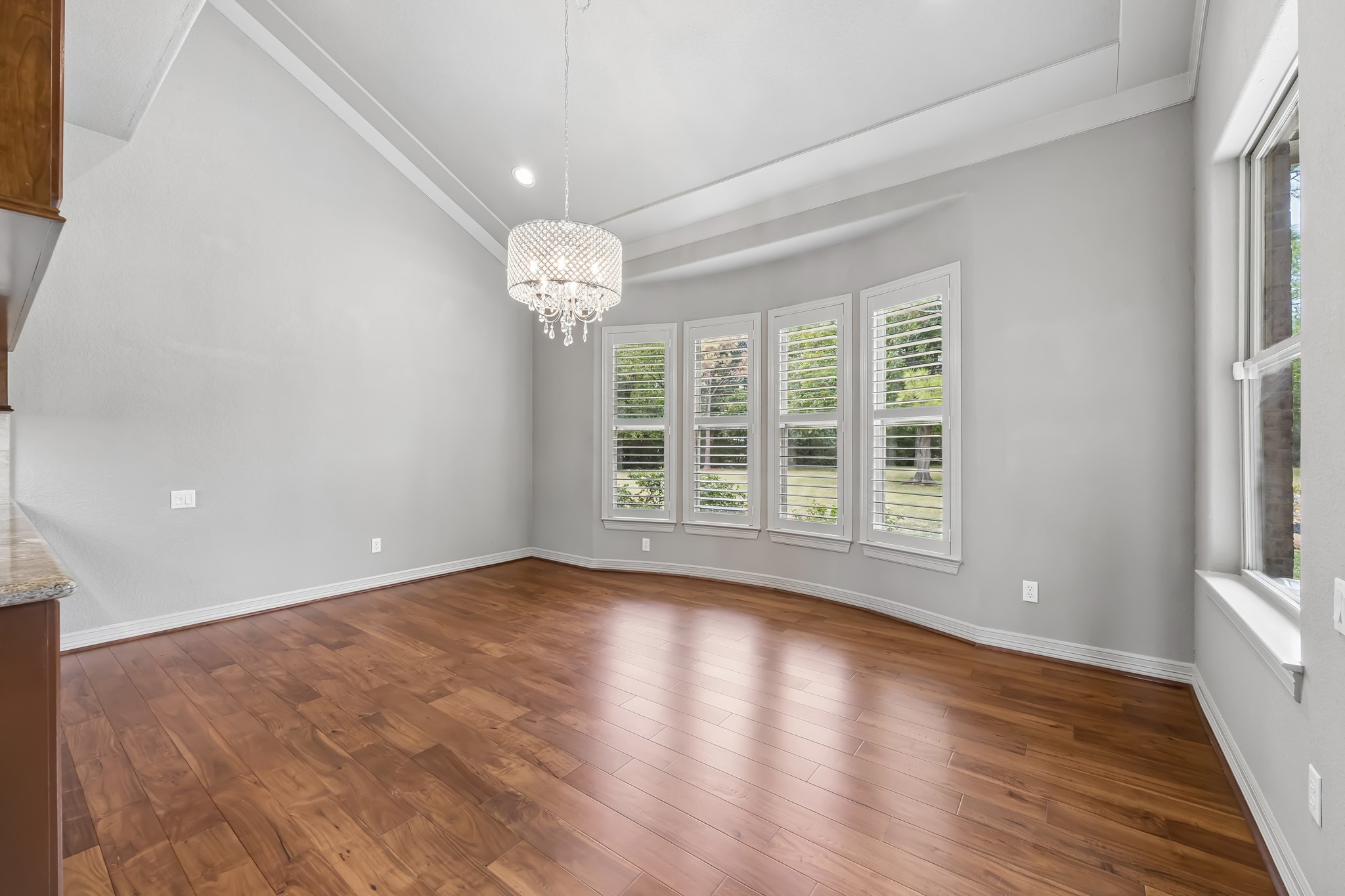 Elegant dining area featuring rich hardwood floors, a stunning chandelier, and large windows with plantation shutters overlooking the serene outdoors.