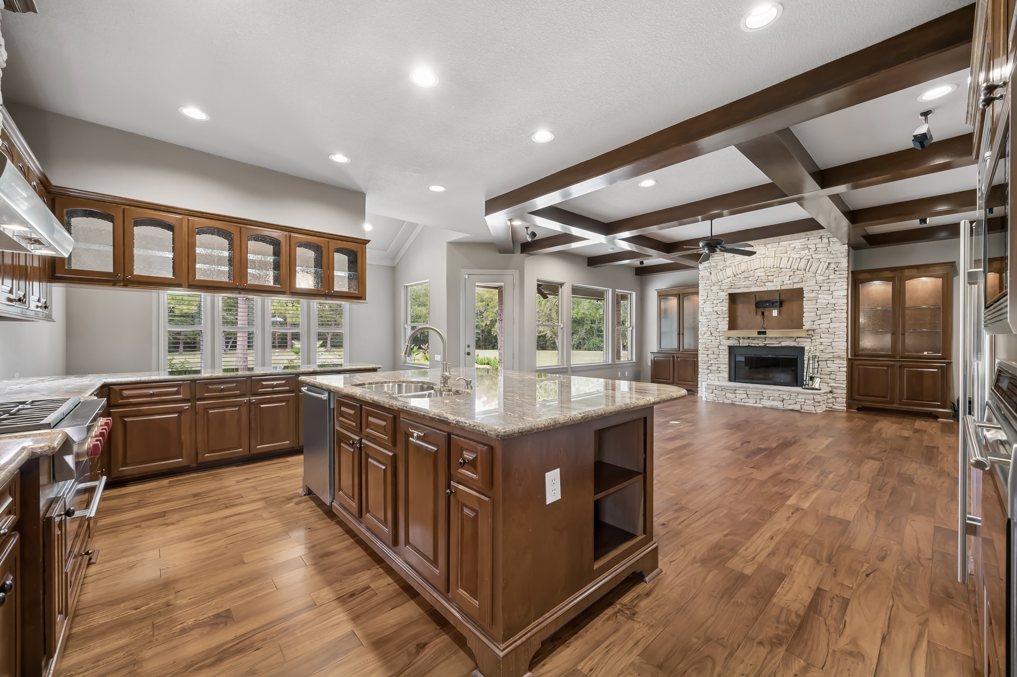Spacious open-concept kitchen and living area featuring rich wooden cabinetry, a large central island with granite countertops, and a cozy stone fireplace, complemented by elegant ceiling beams and abundant natural light.