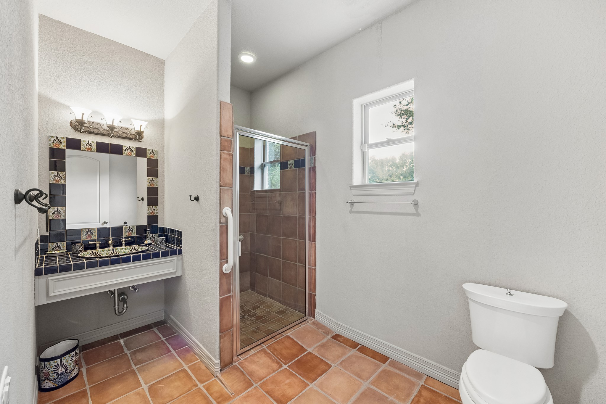 Charming bathroom featuring a tiled walk-in shower, elegant vanity with decorative backsplash, and natural light through a small window, all set on rustic terracotta flooring.  Wheel Chair accessable