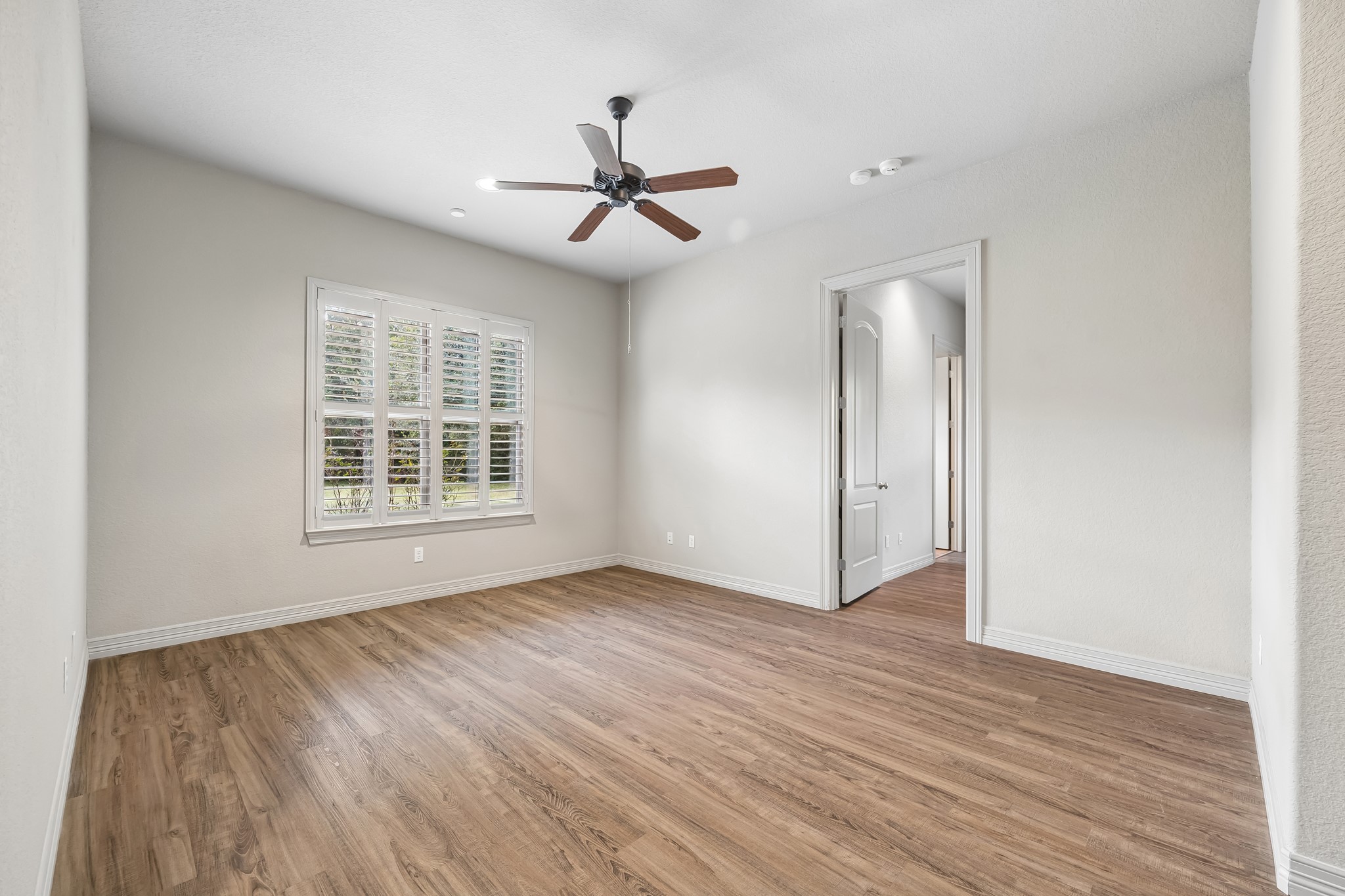 Bright and airy room featuring elegant wood flooring, a ceiling fan, and large window with plantation shutters, offering a serene view and abundant natural light. Guest living area.