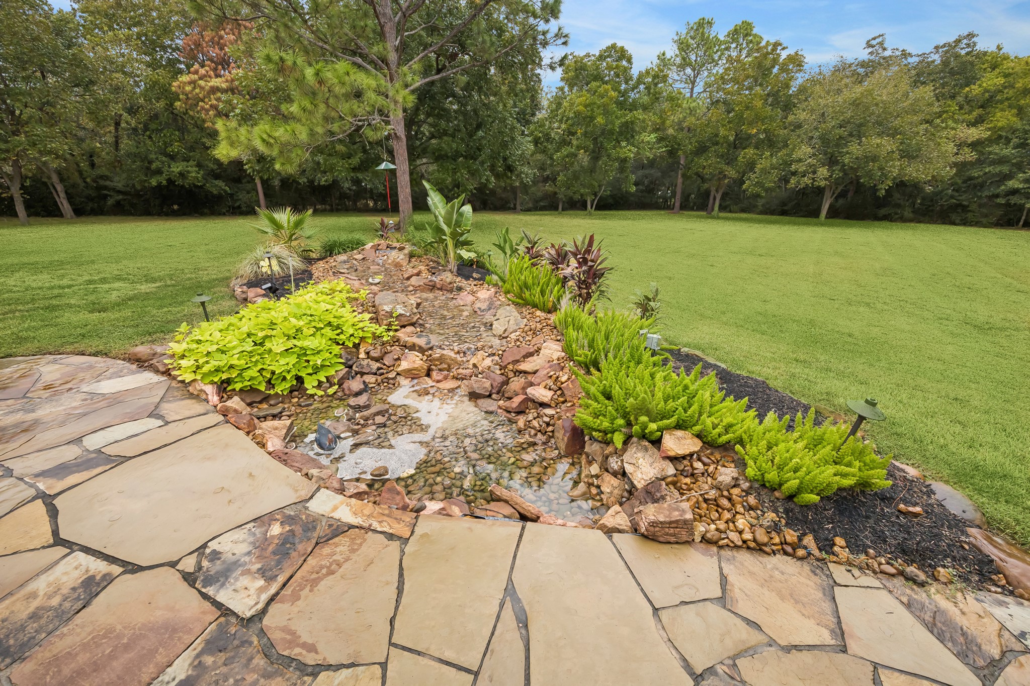 Discover a serene backyard oasis featuring a beautifully landscaped garden with a tranquil rock-lined water feature, bordered by lush greenery and a spacious lawn.