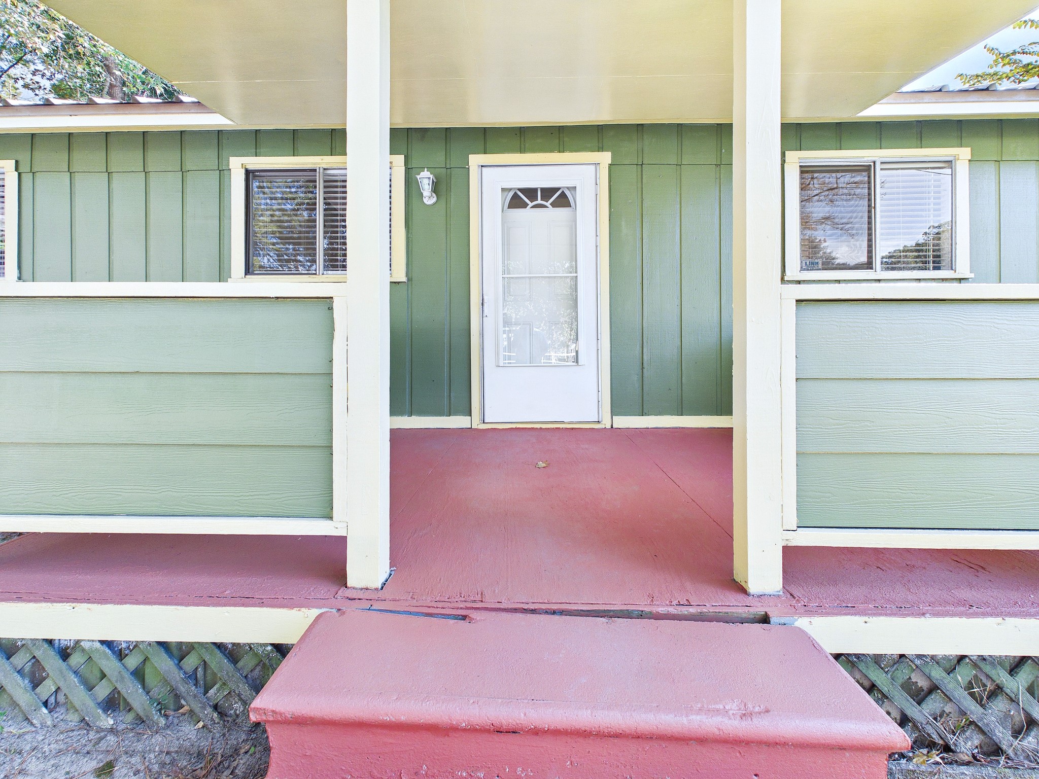 Charming home entrance with a welcoming covered porch, featuring vibrant green siding and contrasting red flooring, perfect for relaxing outdoors.