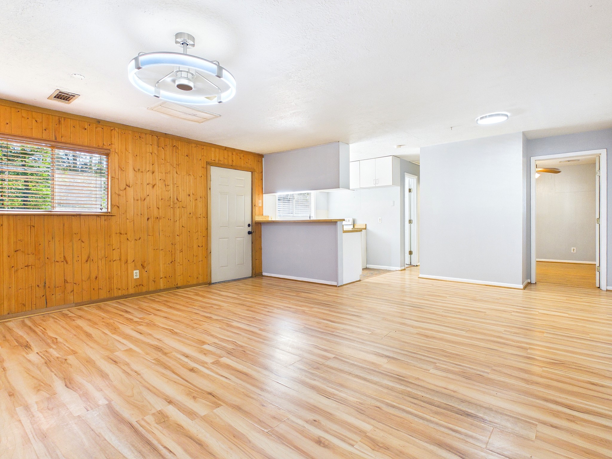 Spacious open-concept living area with warm wood paneling, sleek wood-look floors, and modern lighting, seamlessly connected to a cozy kitchen.