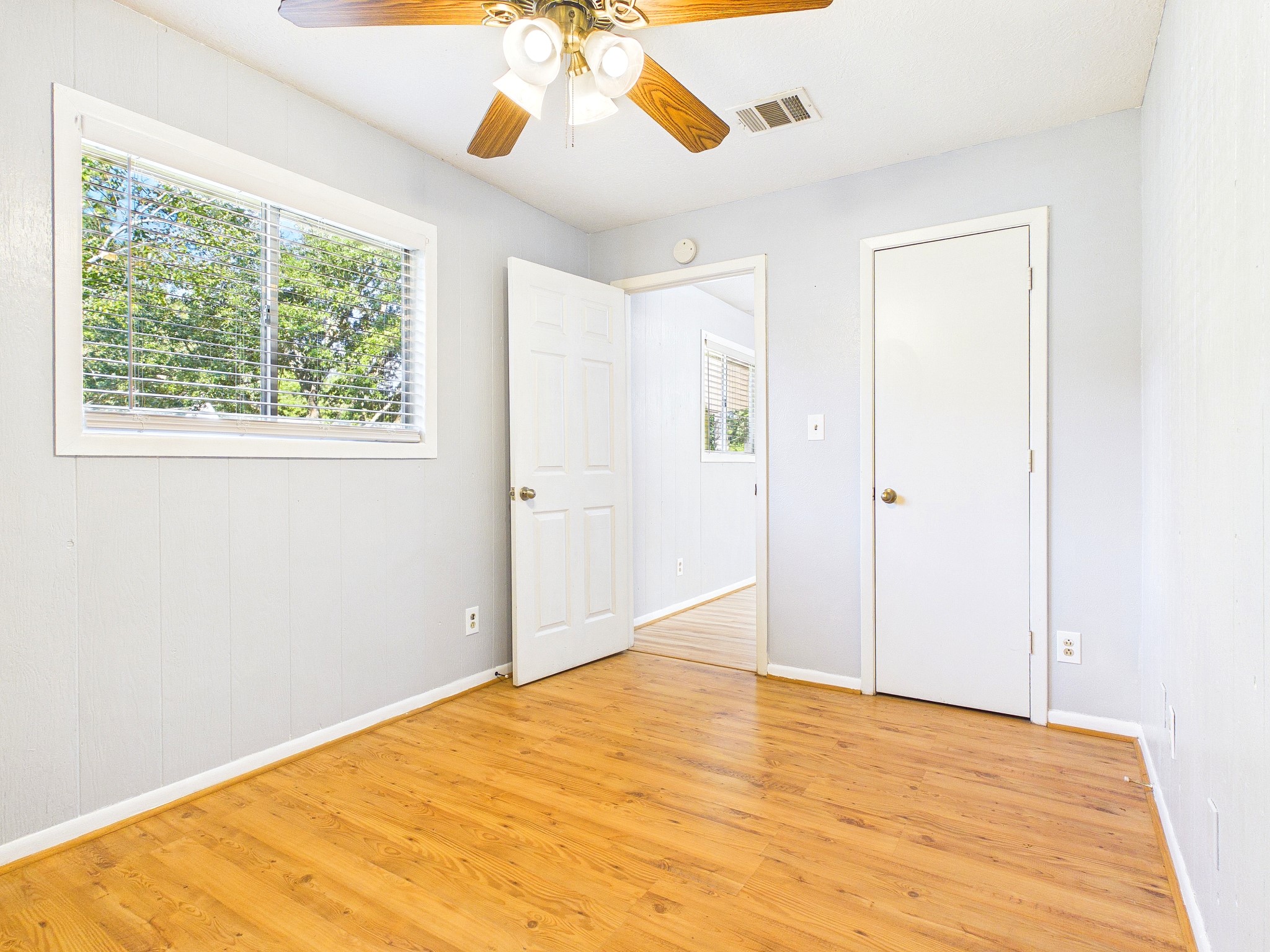 Bright bedroom with wood-look floors, a ceiling fan, and ample natural light from a large window.