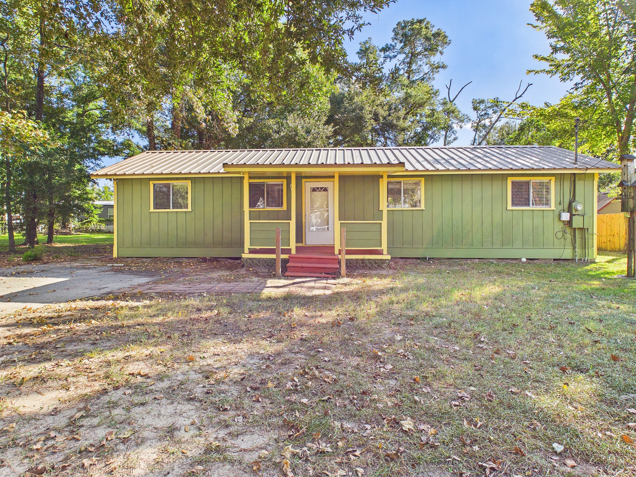 Charming single-story home with a distinctive green exterior, metal roof, and inviting front porch, nestled among mature trees in a serene setting.