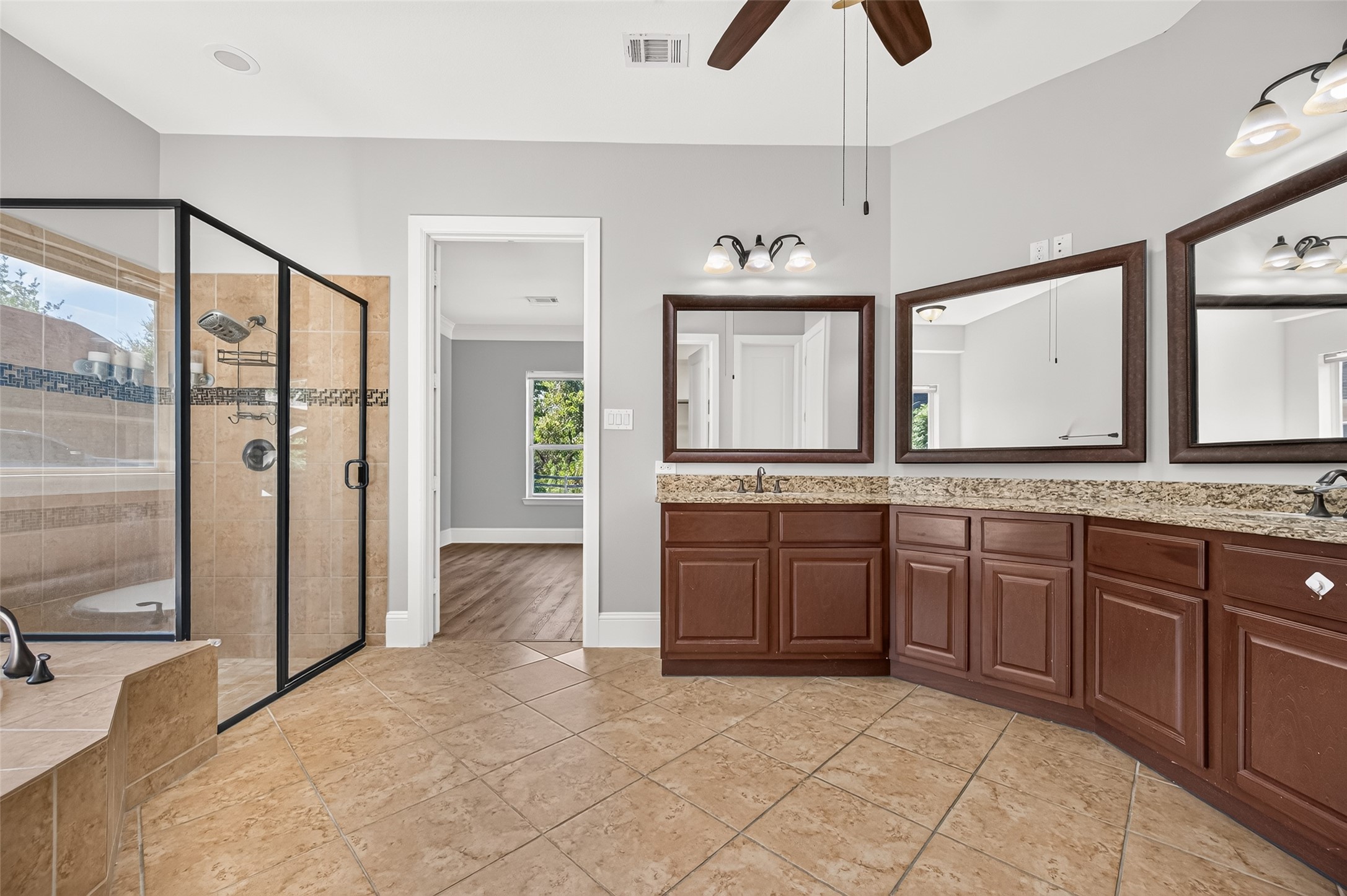 Spa-like primary bath! Indulge in the deep corner soaking tub with elegant tile detailing. Features a massive granite-topped vanity with dual sinks, rich wood cabinetry, a large framed mirror, stylish lighting, and a massive walk-in layout.