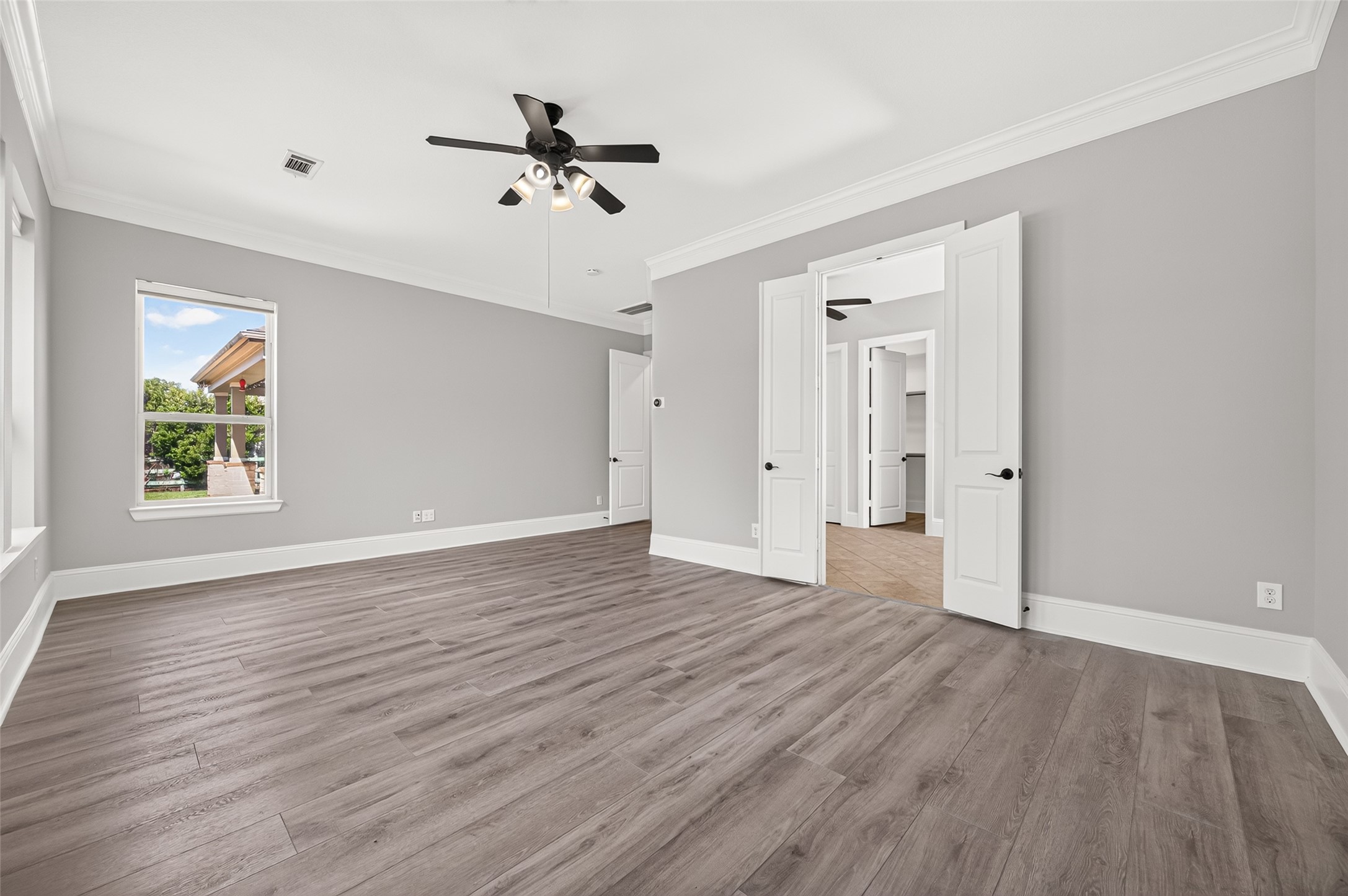 Serene primary suite retreat! This spacious bedroom boasts stylish, modern wood-look flooring, elegant crown molding, a classic ceiling fan, and a wall of large windows overlooking the lush, private backyard space.
