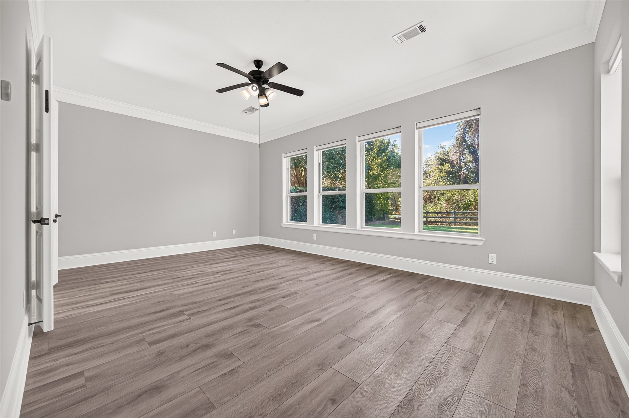 Serene primary suite retreat! This spacious bedroom boasts stylish, modern wood-look flooring, elegant crown molding, a classic ceiling fan, and a wall of large windows overlooking the lush, private backyard space.