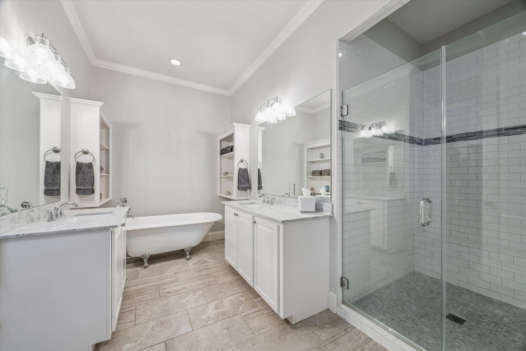 This bathroom features a clean and elegant design with a glass-enclosed walk-in shower, a classic clawfoot bathtub and dual vanities with ample counter space and storage