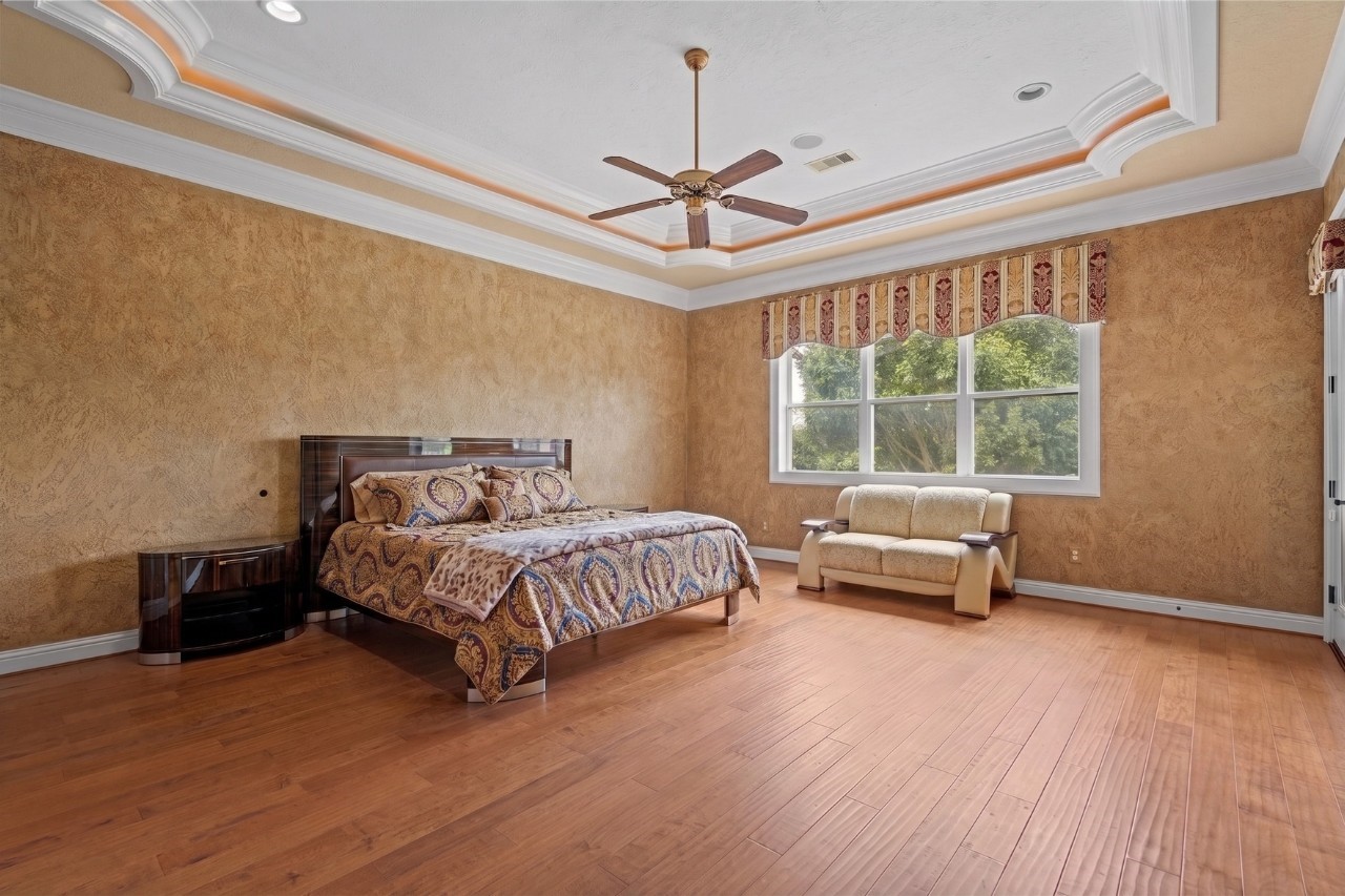 Spacious secondary suite upstairs showcasing an ornate illuminated tray ceiling, rich hardwood flooring, and large windows with serene treetop views.