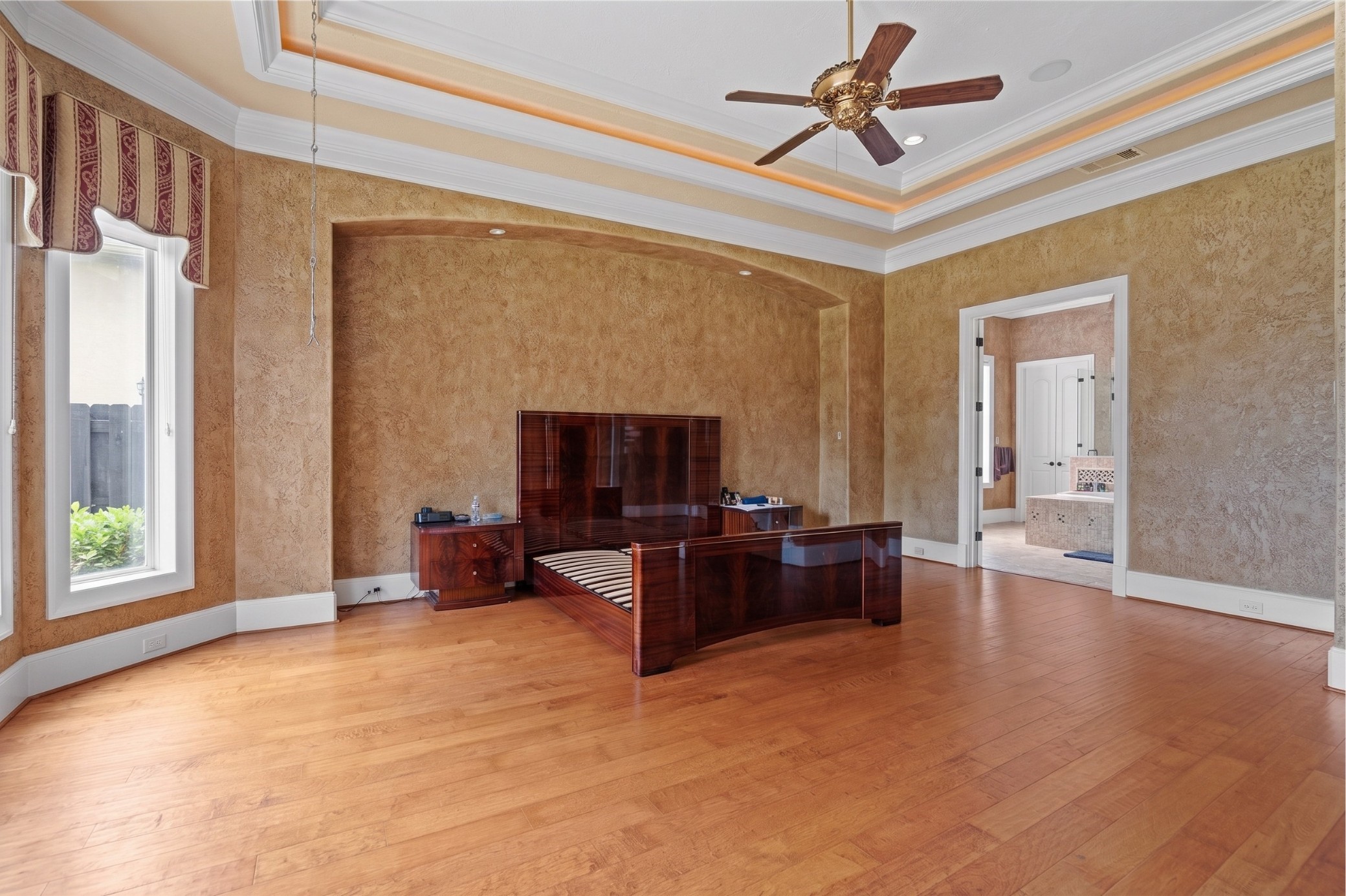 Expansive primary suite highlights an illuminated tray ceiling, warm hardwood flooring, a graceful arched alcove, and seamless access to the luxurious bath.