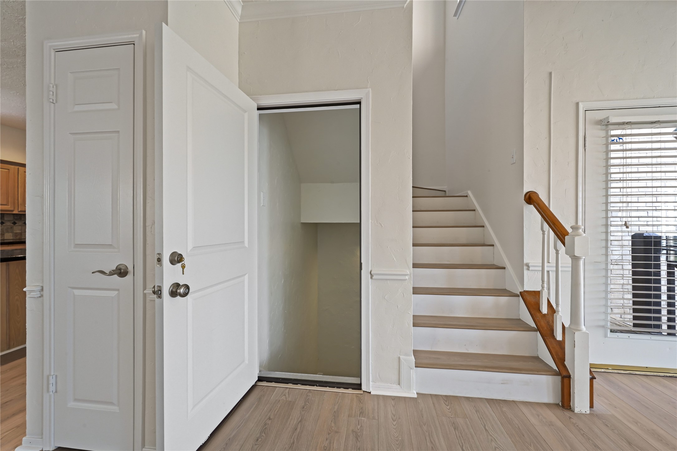 A well-lit entryway features a staircase with elegant wooden handrails, a convenient storage closet, and a view into a bright kitchen area, enhancing the home's welcoming appeal.