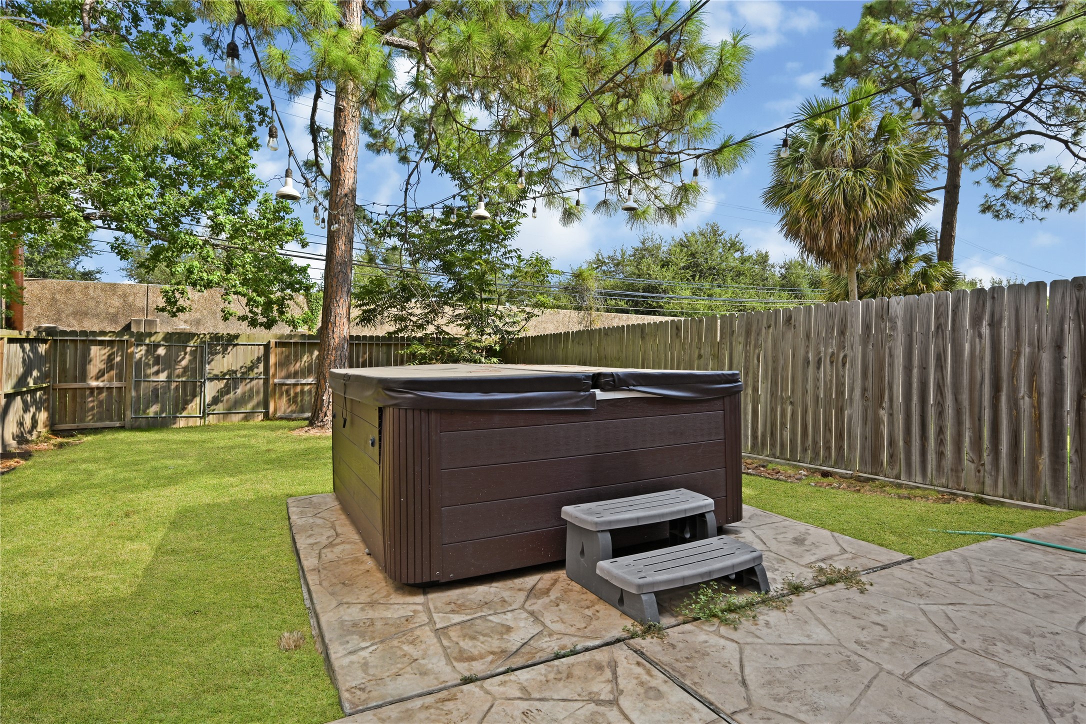 Experience relaxation in this private backyard featuring a luxurious hot tub, surrounded by lush greenery and a charming wooden fence for ultimate seclusion.