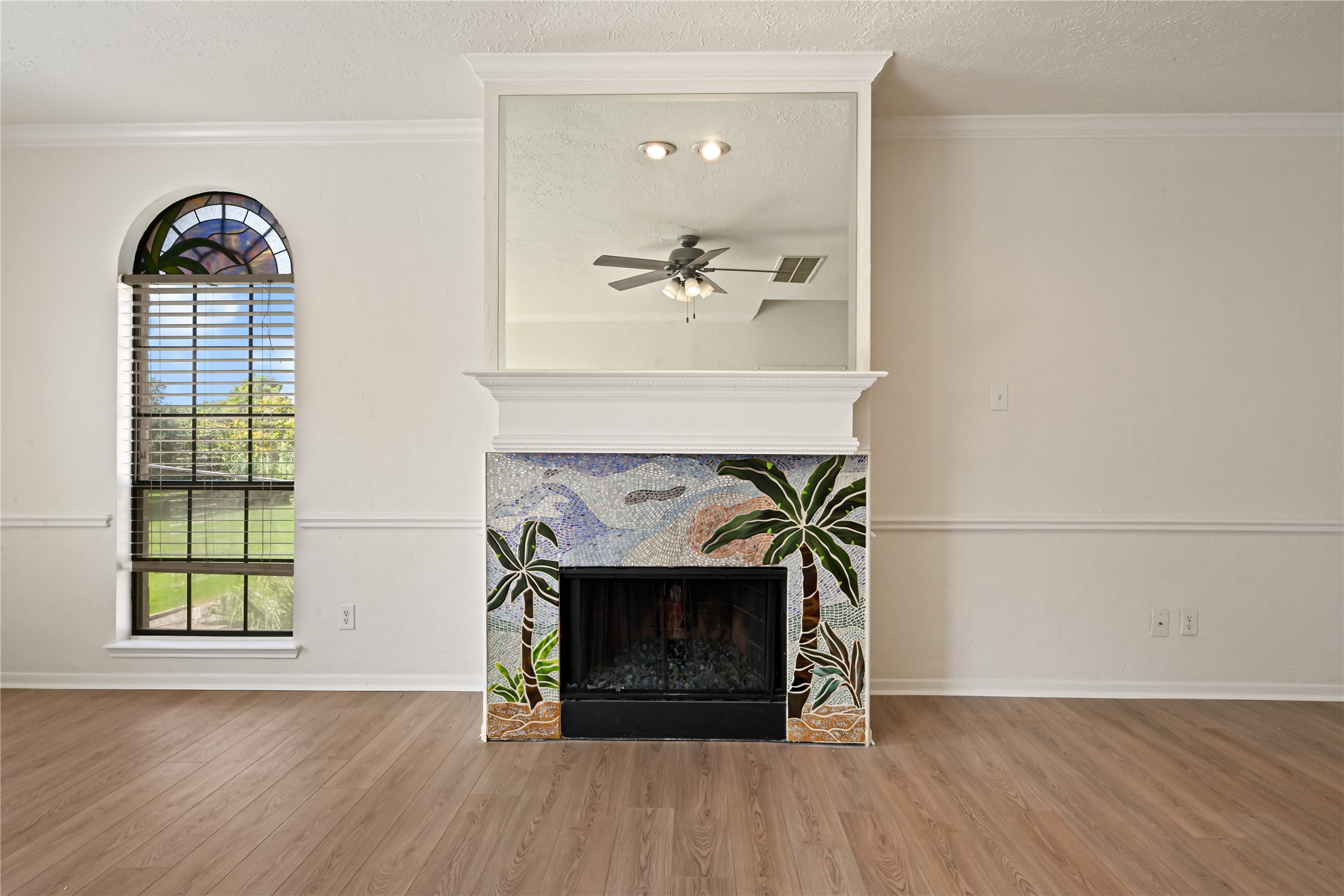 Inviting living space featuring a unique mosaic-tiled fireplace with a tropical motif, complemented by elegant wood flooring and a stylish arched window that lets in abundant natural light.