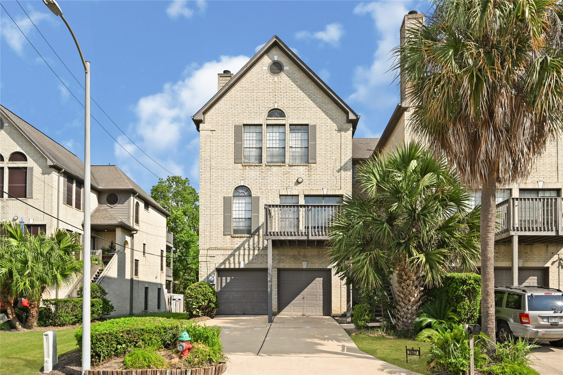 Charming multi-level townhouse with a brick exterior, featuring a spacious two-car garage and lush palm landscaping, set in a serene neighborhood.